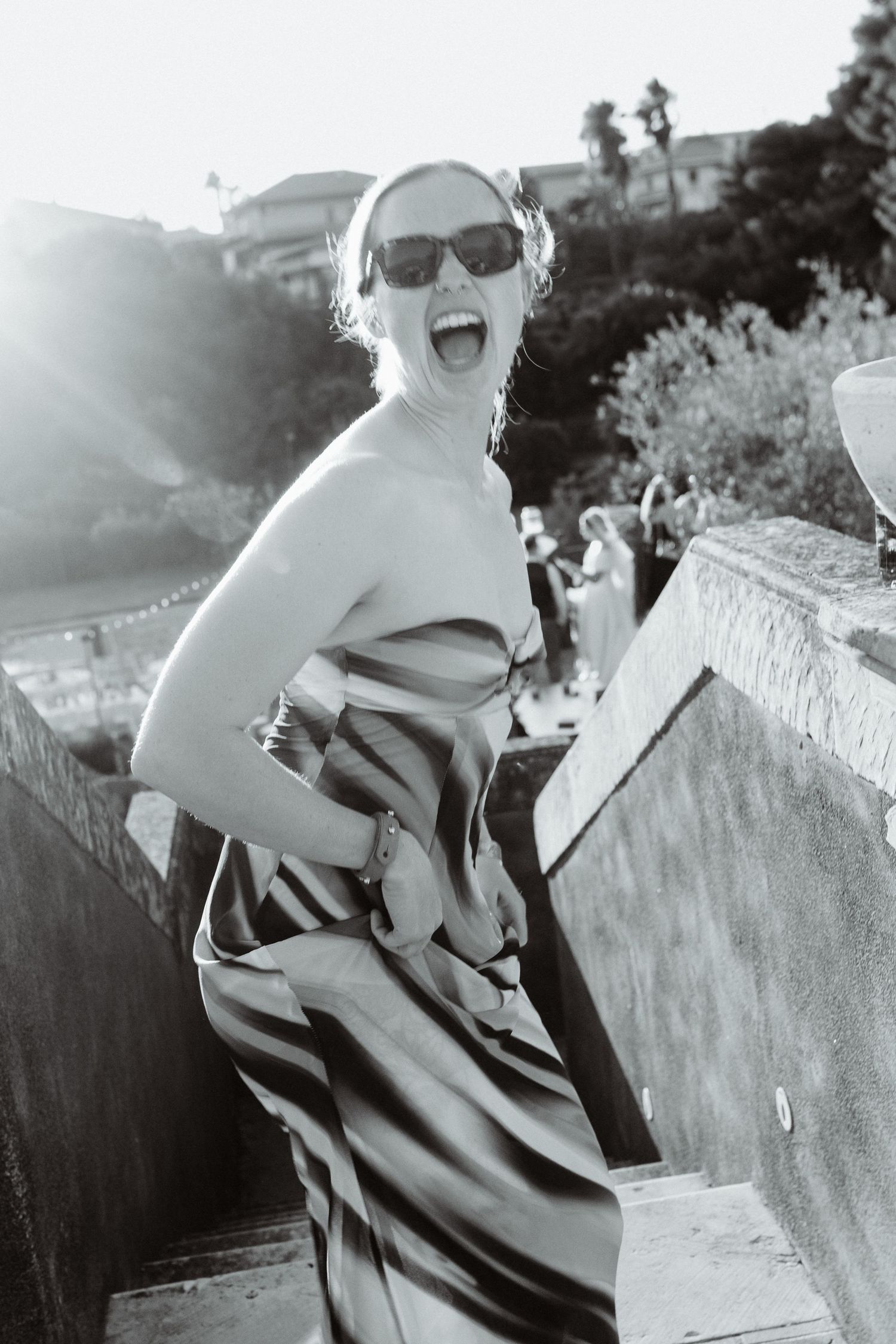 Black and white photo of a laughing woman in sunglasses and strapless striped dress posing on outdoor steps.