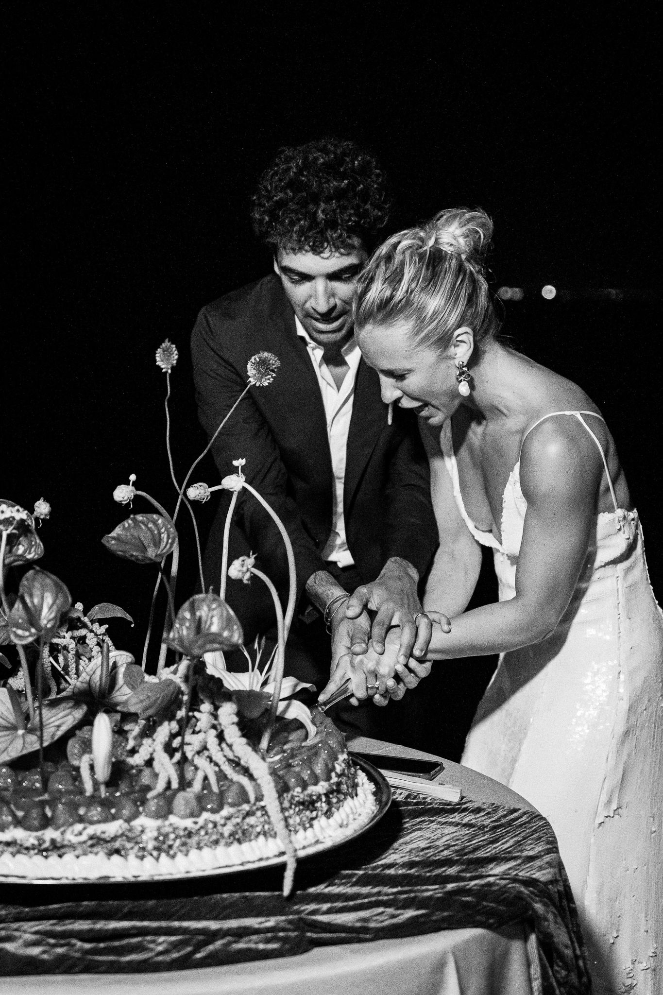 Newlyweds working together to cut their decorative wedding cake in dramatic black and white.