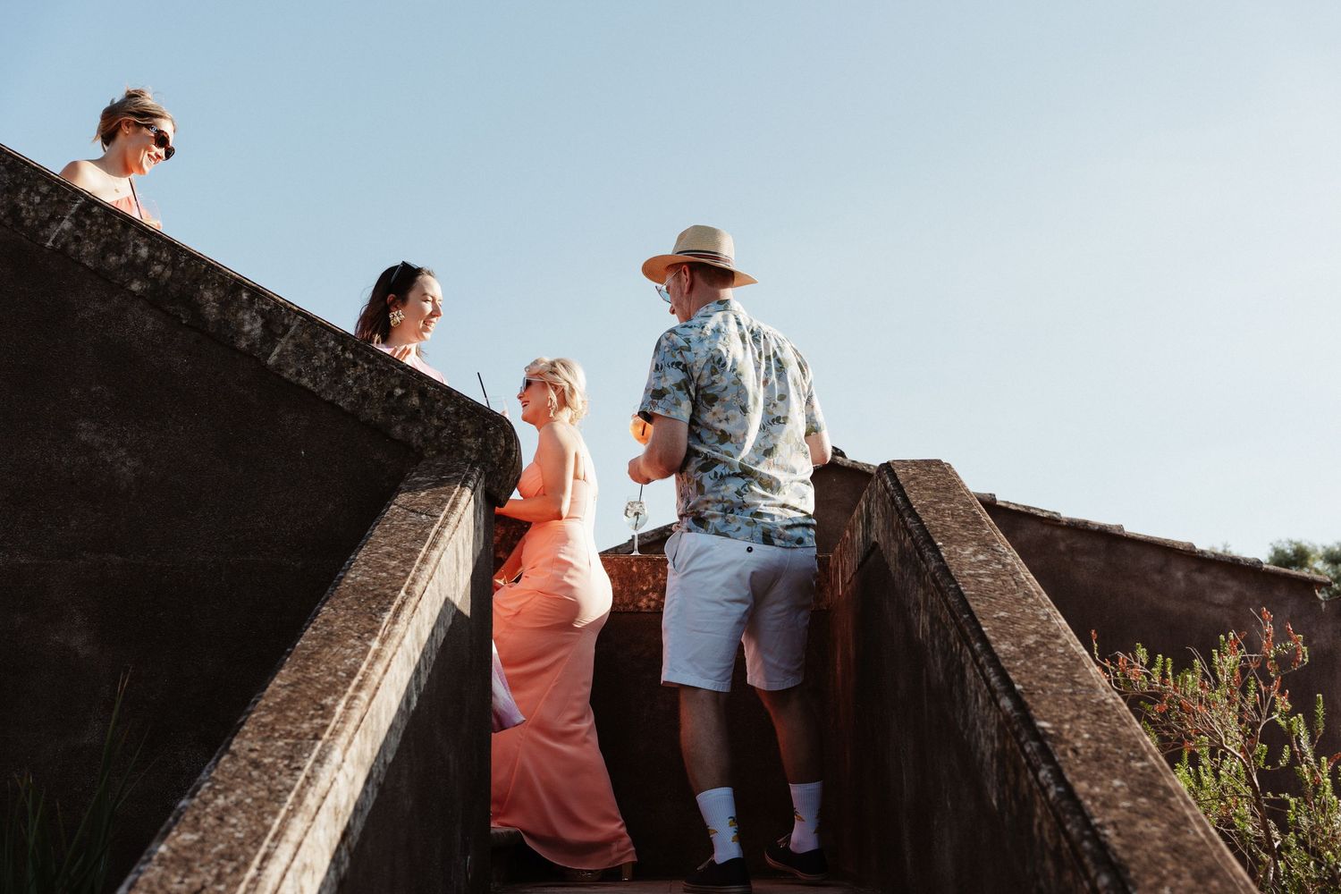 Group of people in summer attire conversing on stone steps against a clear blue sky.