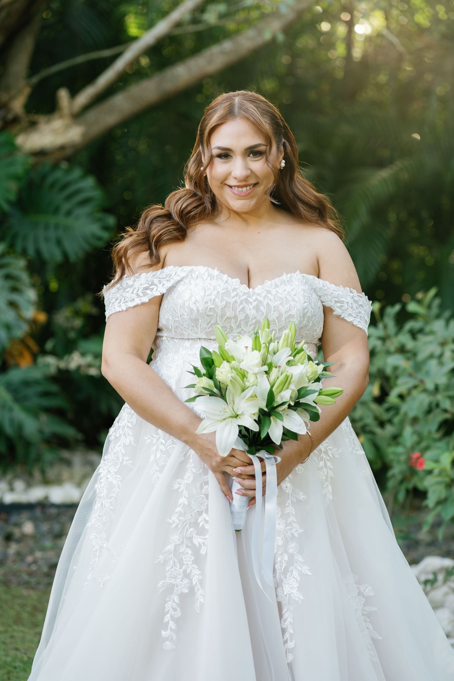 Smiling bride in off-shoulder lace wedding gown holding white lily bouquet outdoors in lush garden setting.