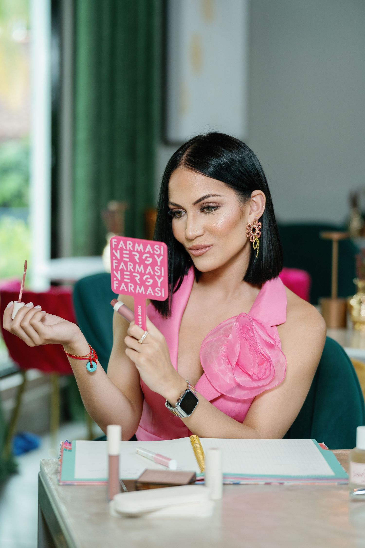 A woman in a pink one-shoulder top holds up a pink decorative sign while sitting at a desk with office supplies.