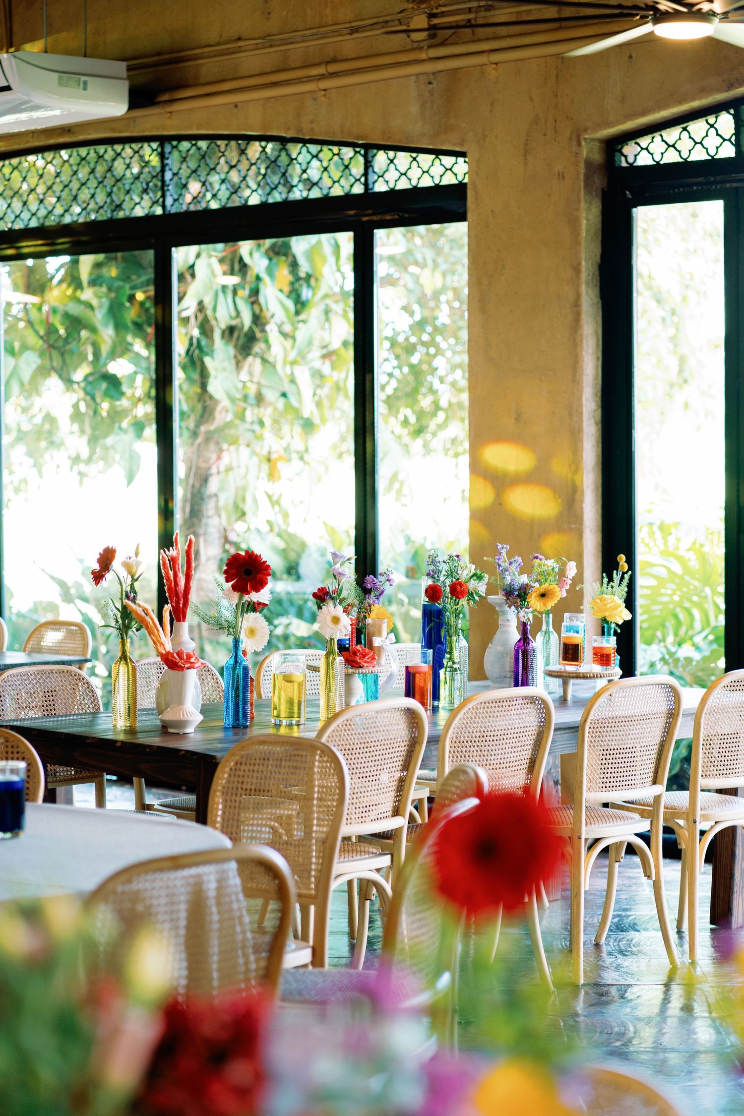 Bright sunlit restaurant interior with colorful floral accents and white rattan chairs.