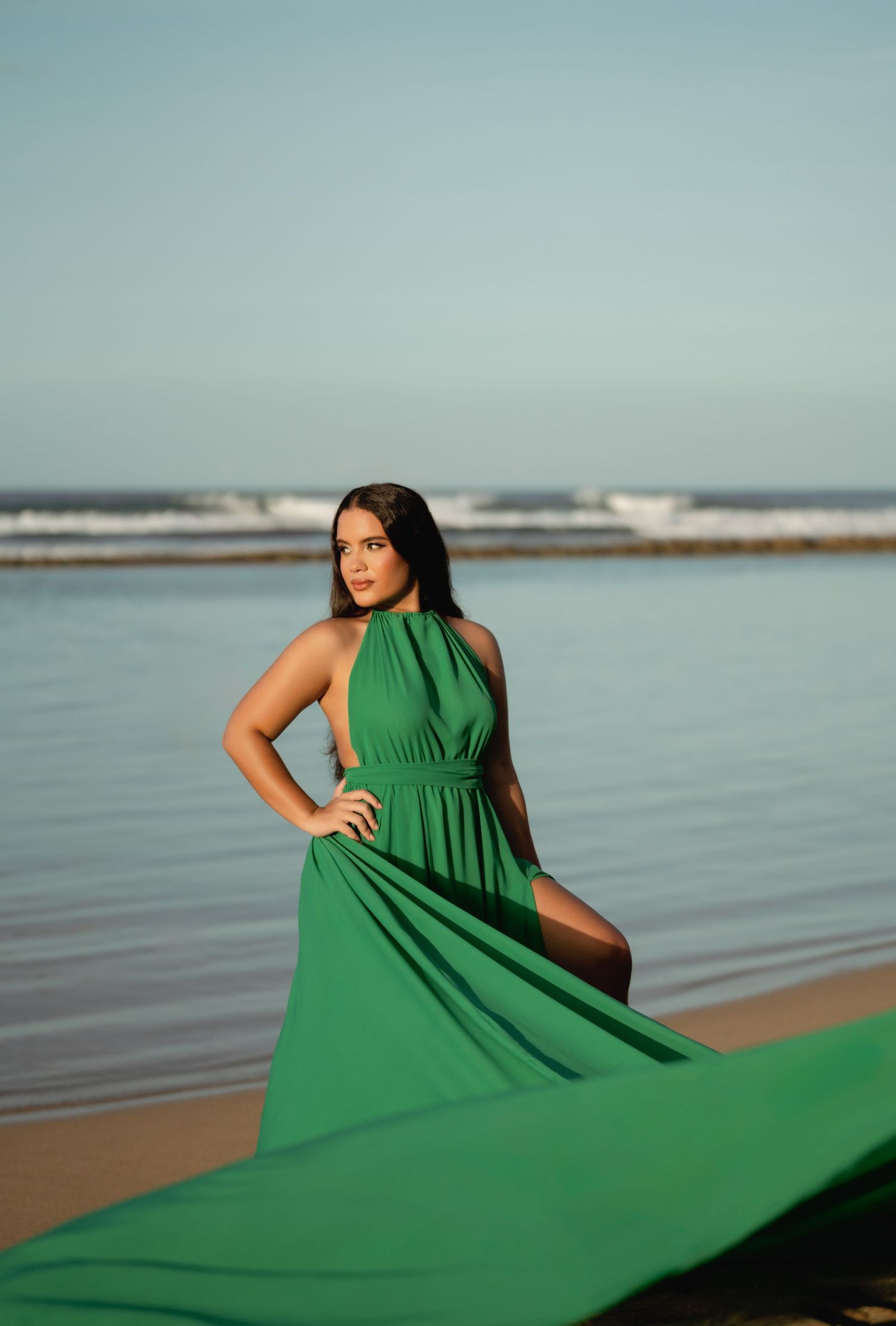 Woman in flowing green dress posing dramatically by ocean at sunset.