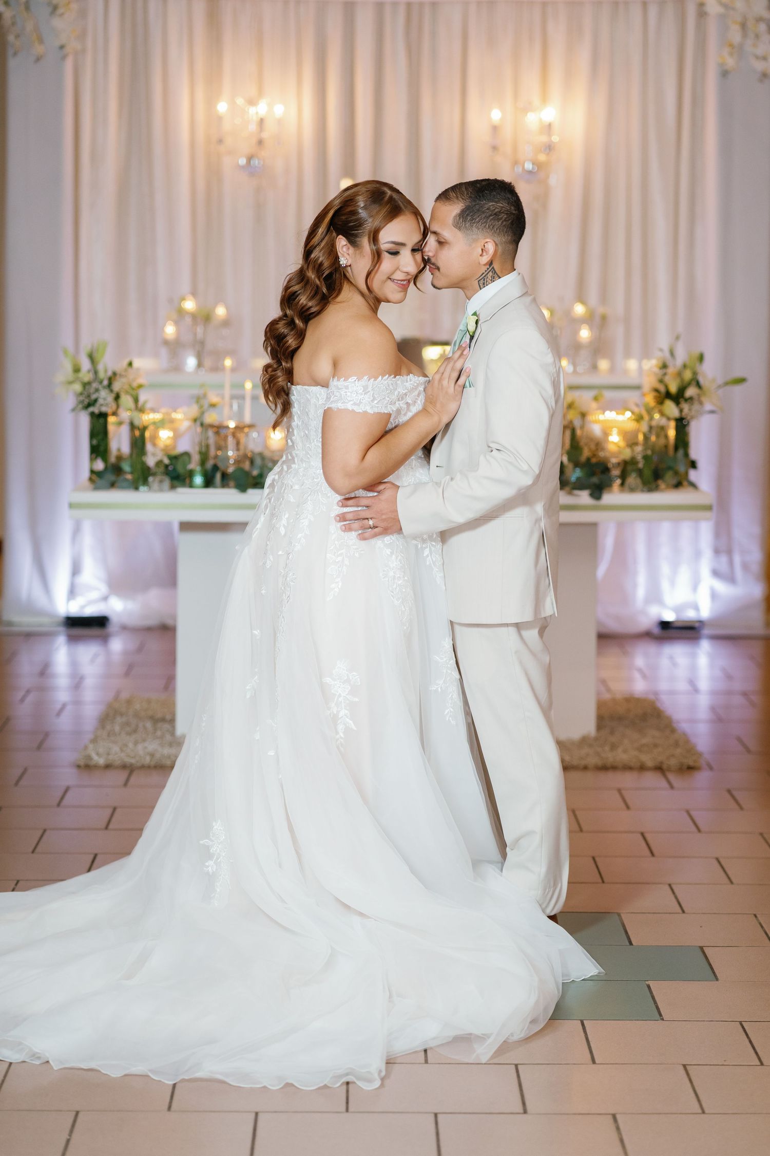 Bride in off-shoulder gown and groom in ivory suit share tender moment on dance floor with candlelit altar backdrop.