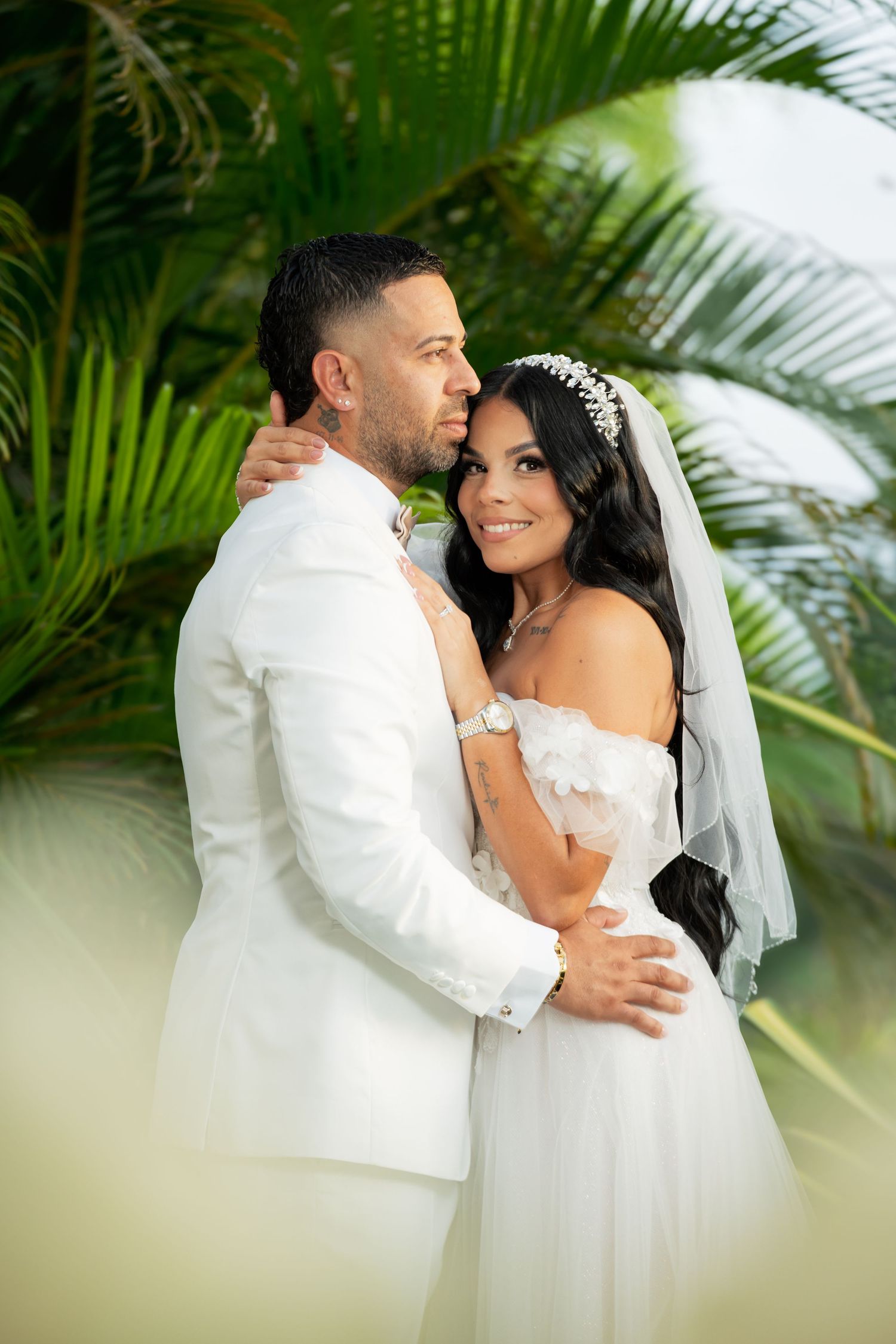 A bride and groom embrace outdoors surrounded by lush tropical palm trees, both dressed in elegant white wedding attire.