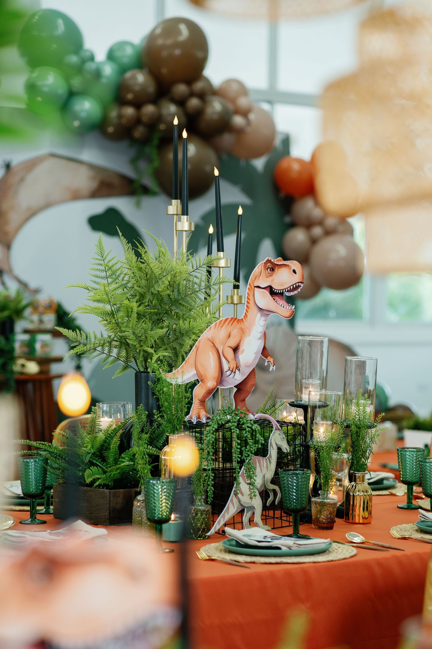 Dinosaur themed table setting with green ferns, vintage glassware, and orange tablecloth.