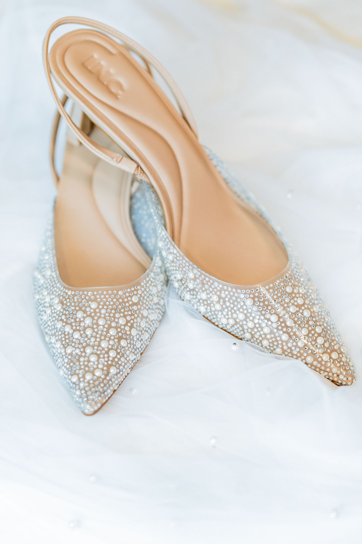 Elegant pearl-embellished slingback wedding heels with pointed toe on white fabric.