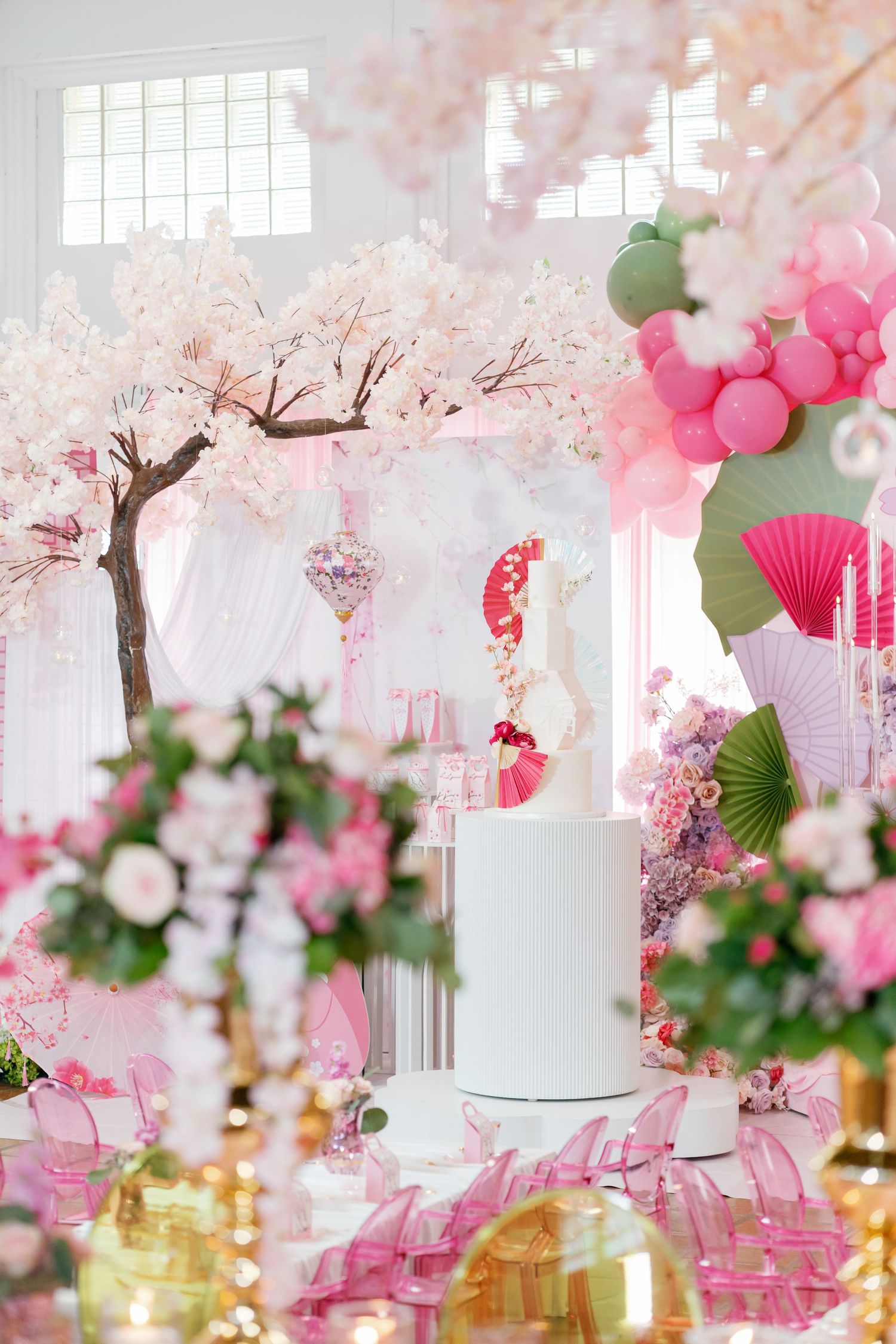 Pink cherry blossom wedding decor with white cake, balloon arch, and floral arrangements in elegant event space.
