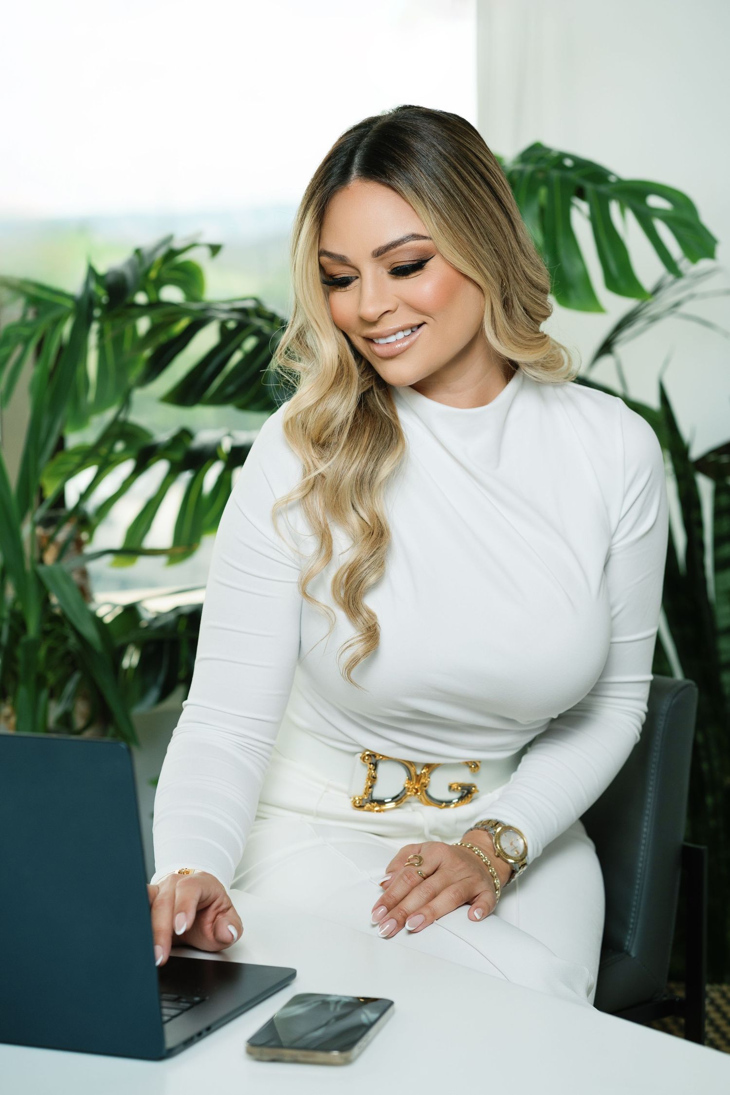 Professional wearing a white long-sleeve dress with designer belt works at desk with laptop and phone near tropical plants.
