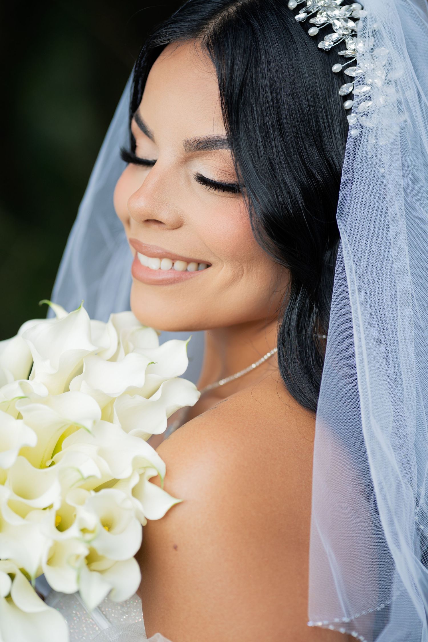 Smiling bride with closed eyes holding white calla lily bouquet, wearing crystal tiara and veil on wedding day.