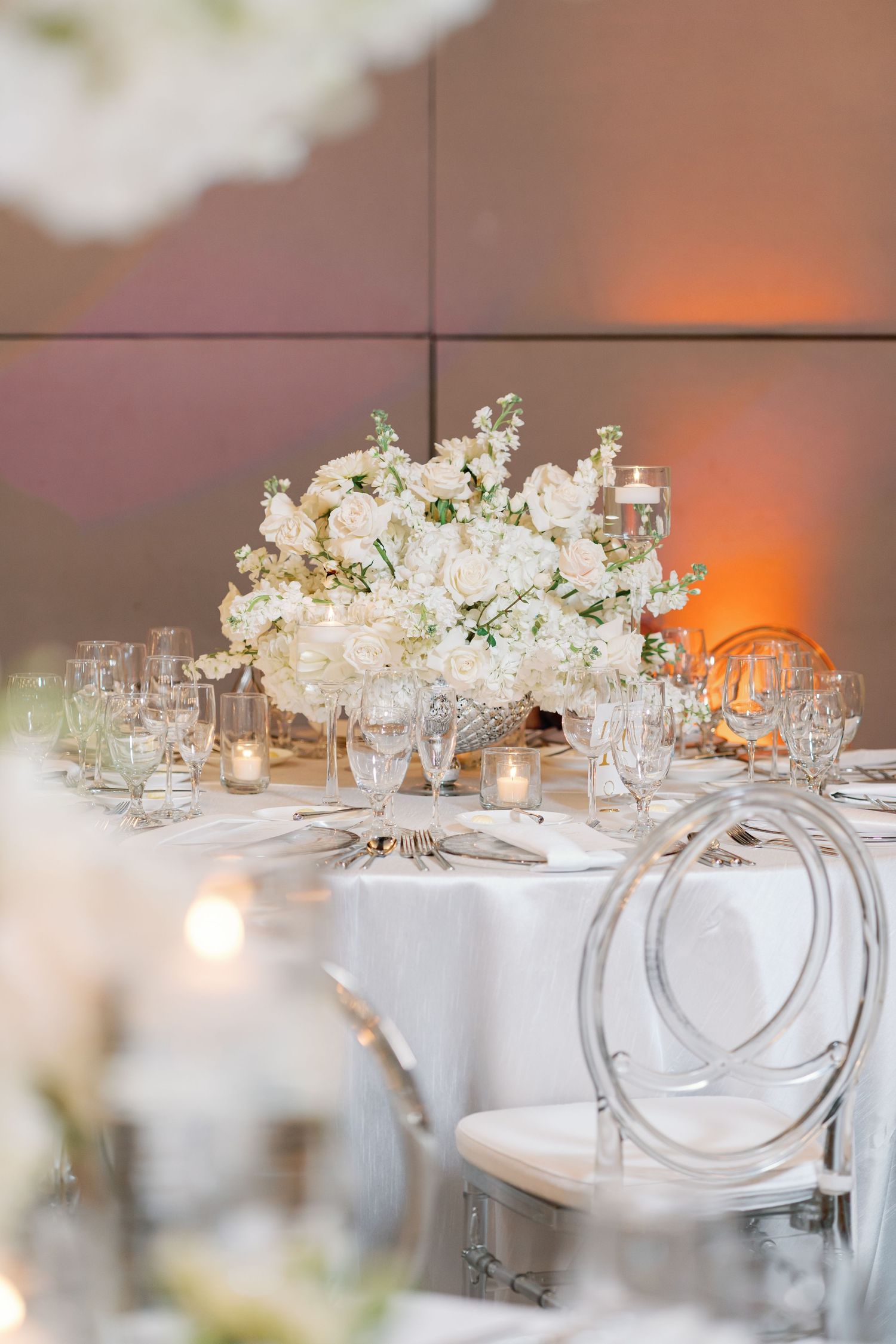Elegant wedding reception table with white floral centerpiece, crystal glassware, candles, and ghost chairs in a softly lit venue.