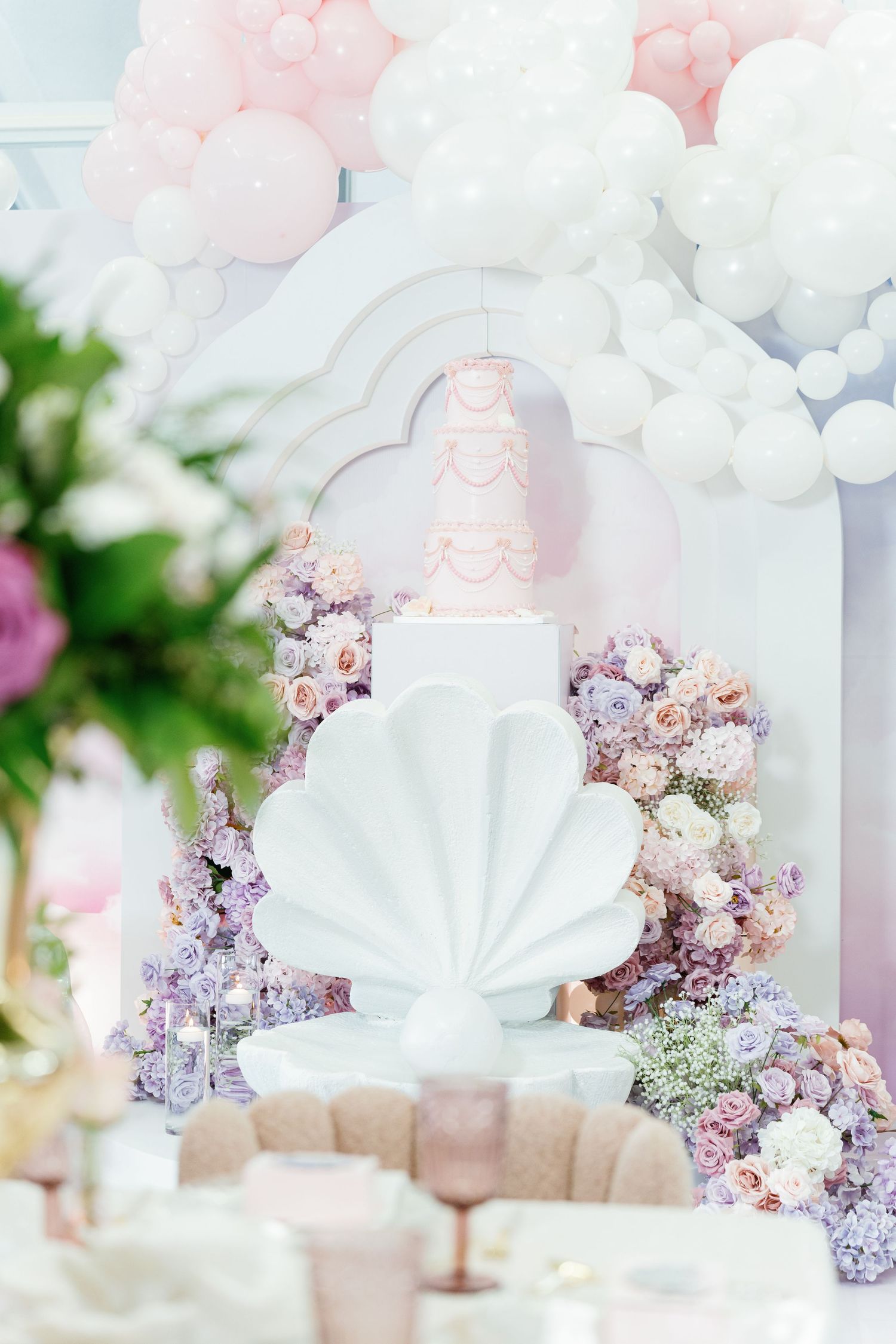 Romantic wedding setup with shell-shaped backdrop, pink balloons, and pastel floral arrangements.