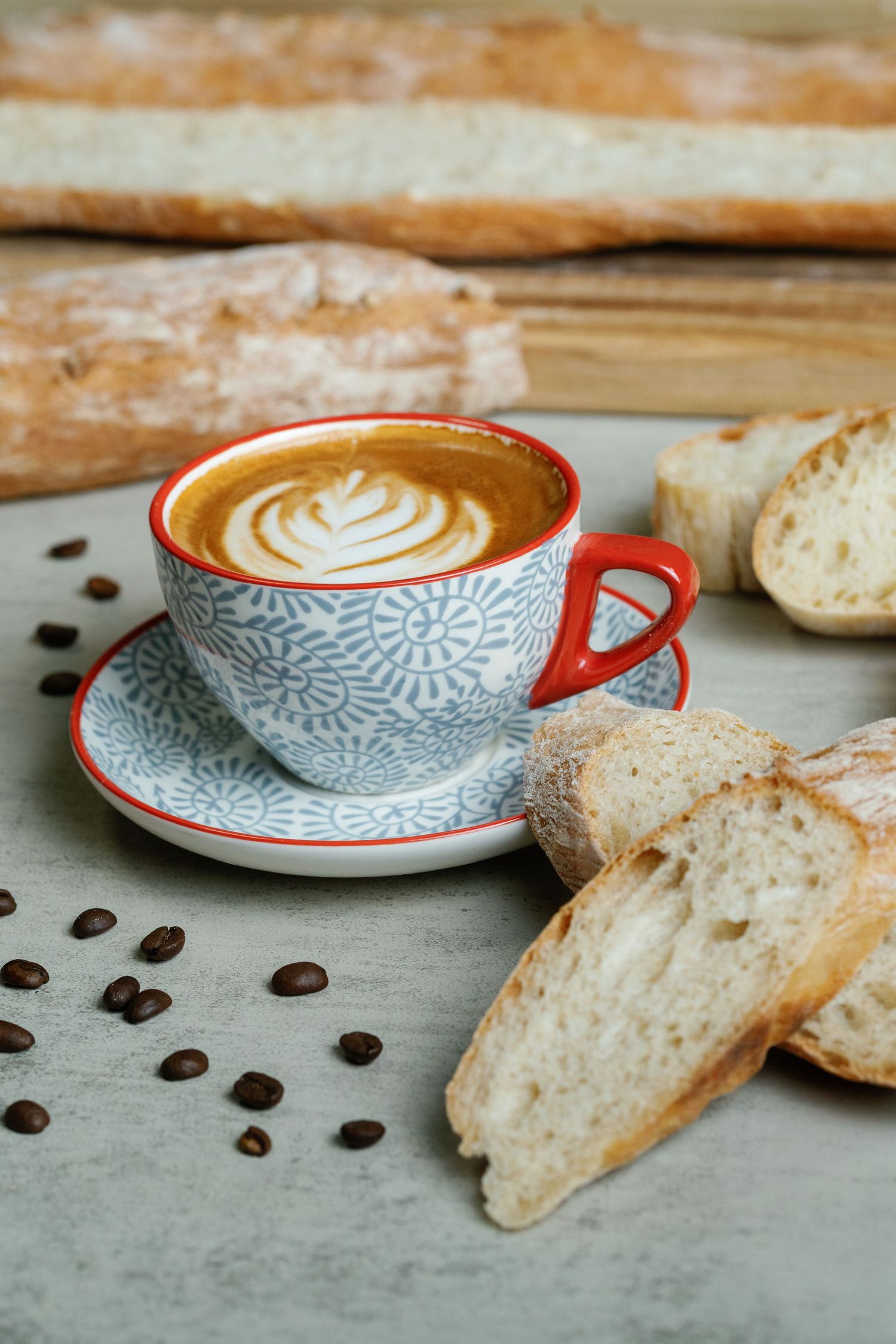 Latte art coffee in red-rimmed cup with sliced bread and scattered coffee beans on gray surface.