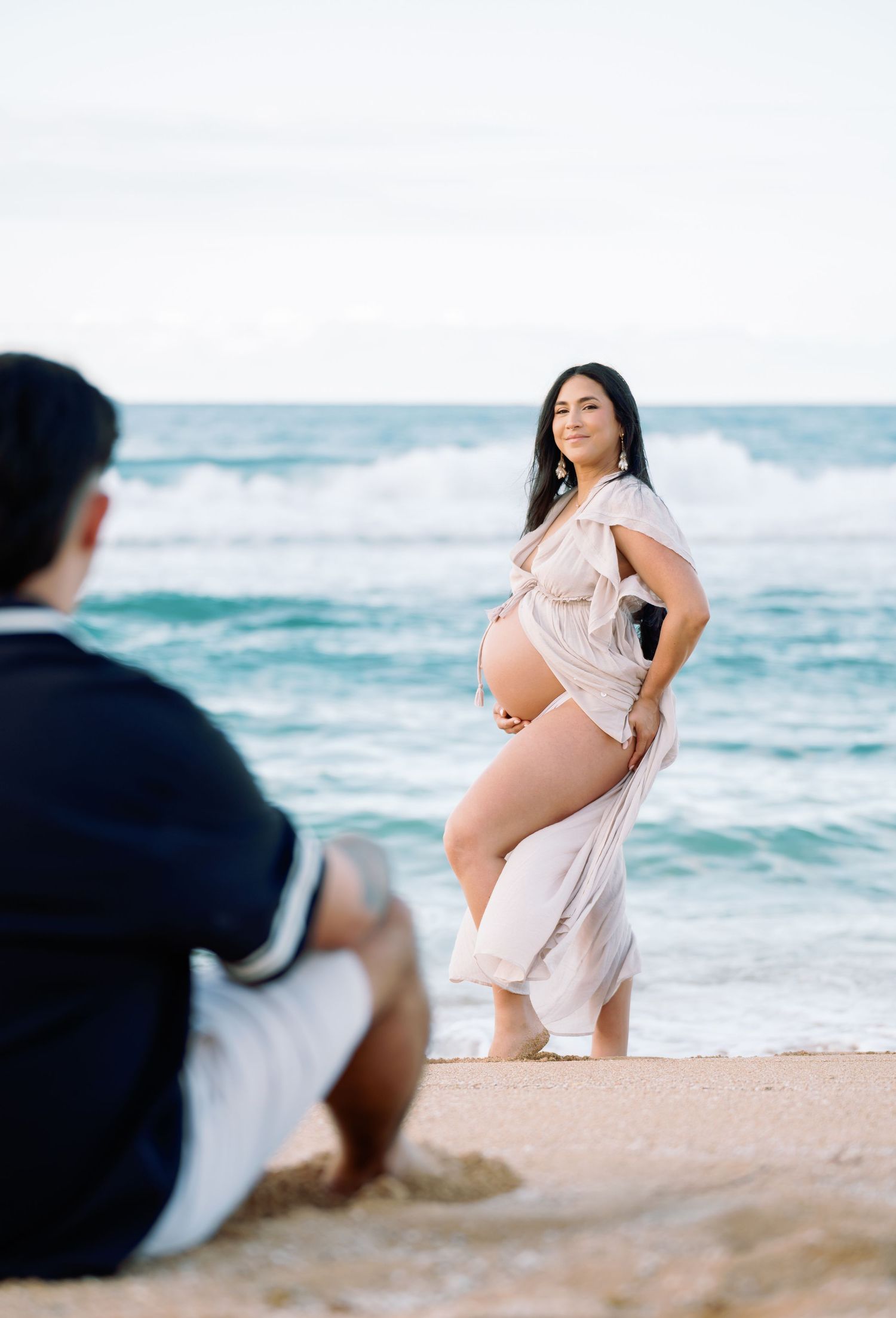 Romantic beach maternity session with flowing white dress against ocean waves.