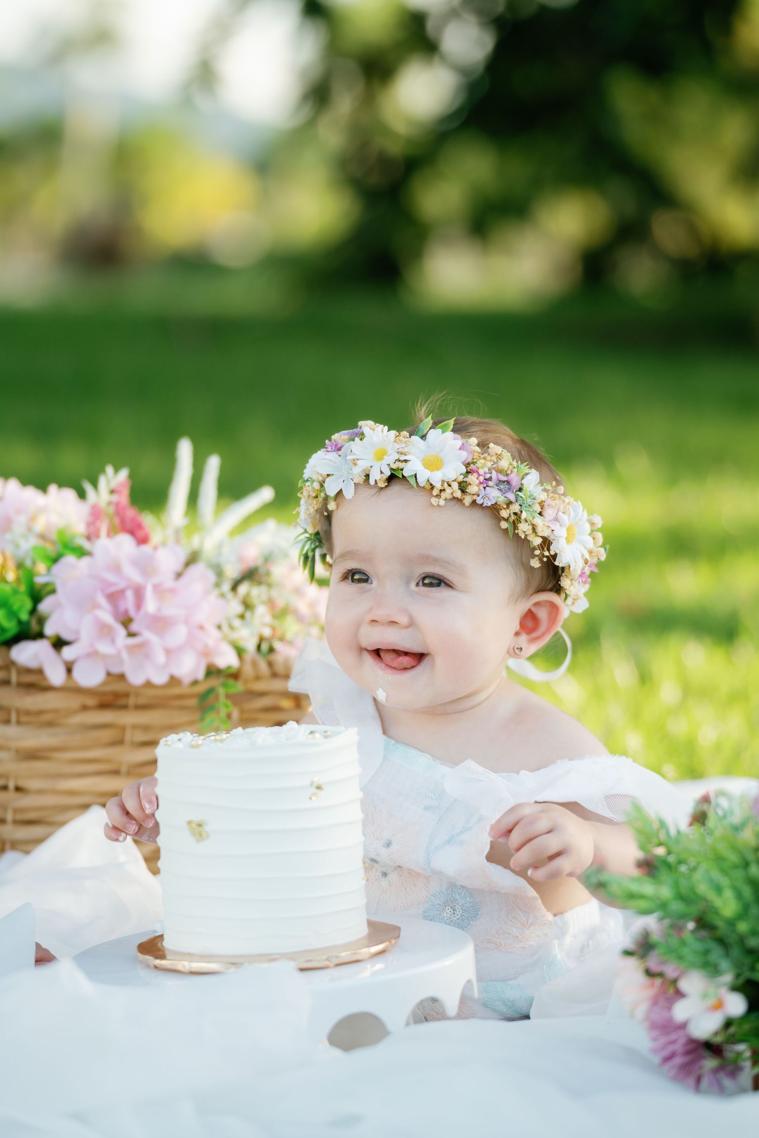 Baby's first birthday celebration with white cake and flower crown in sunny garden setting.