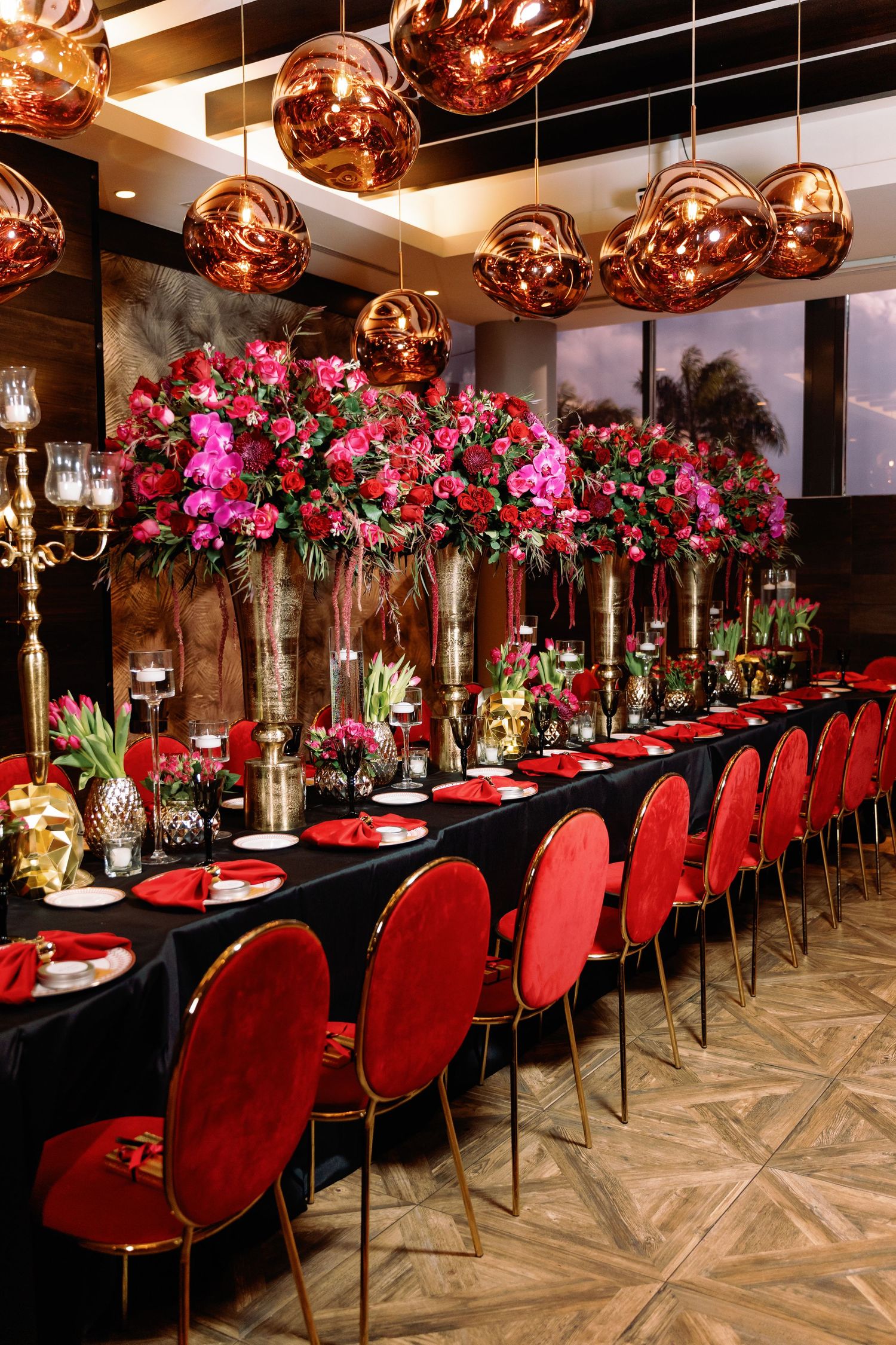 Elegant dining setup with red velvet chairs, copper pendant lights, and vibrant pink floral centerpieces on a dark table.