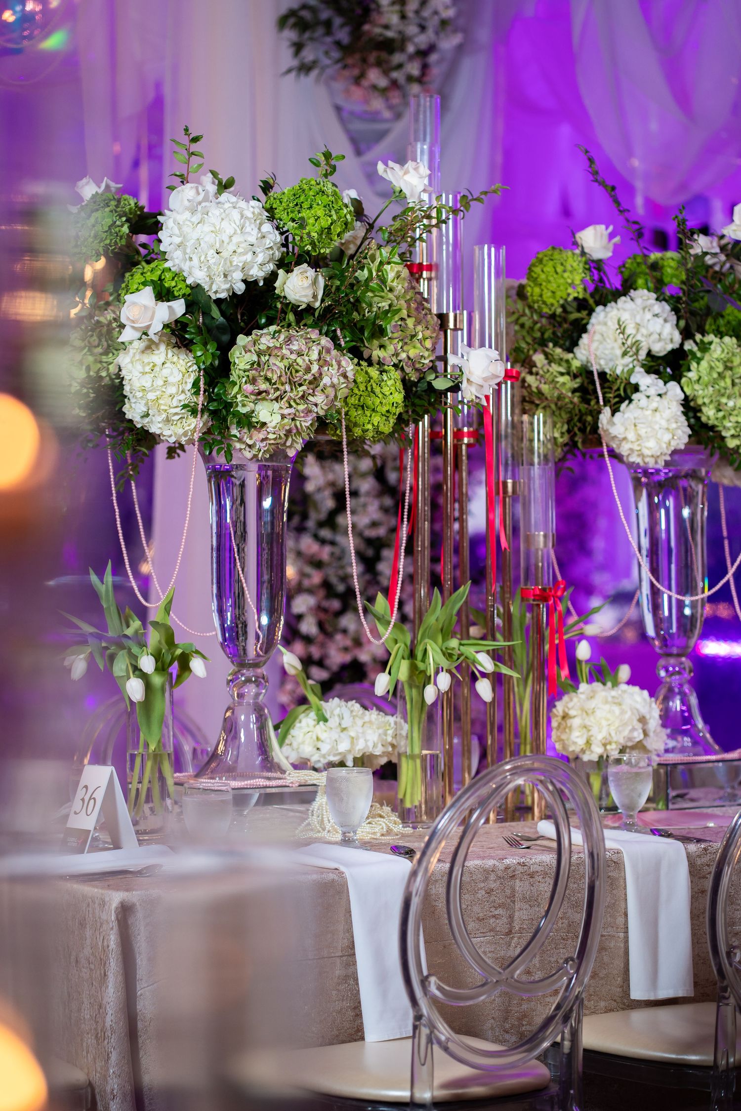 Wedding reception table settings with tall floral arrangements and purple ambient lighting.