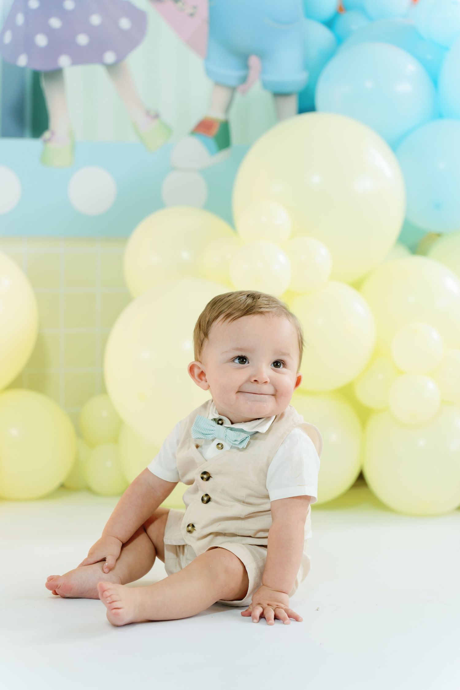 Baby in light colored outfit smiles while lying on floor with pastel blue and yellow balloons in background.
