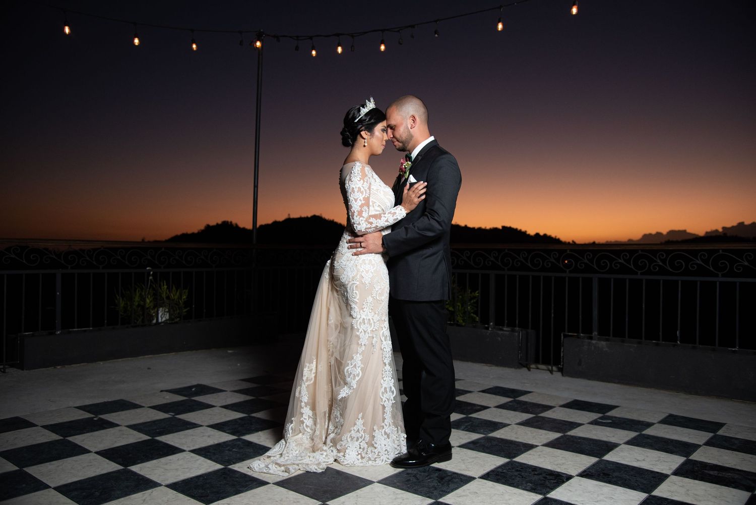 Bride and groom share romantic kiss at sunset on checkered dance floor with string lights overhead.