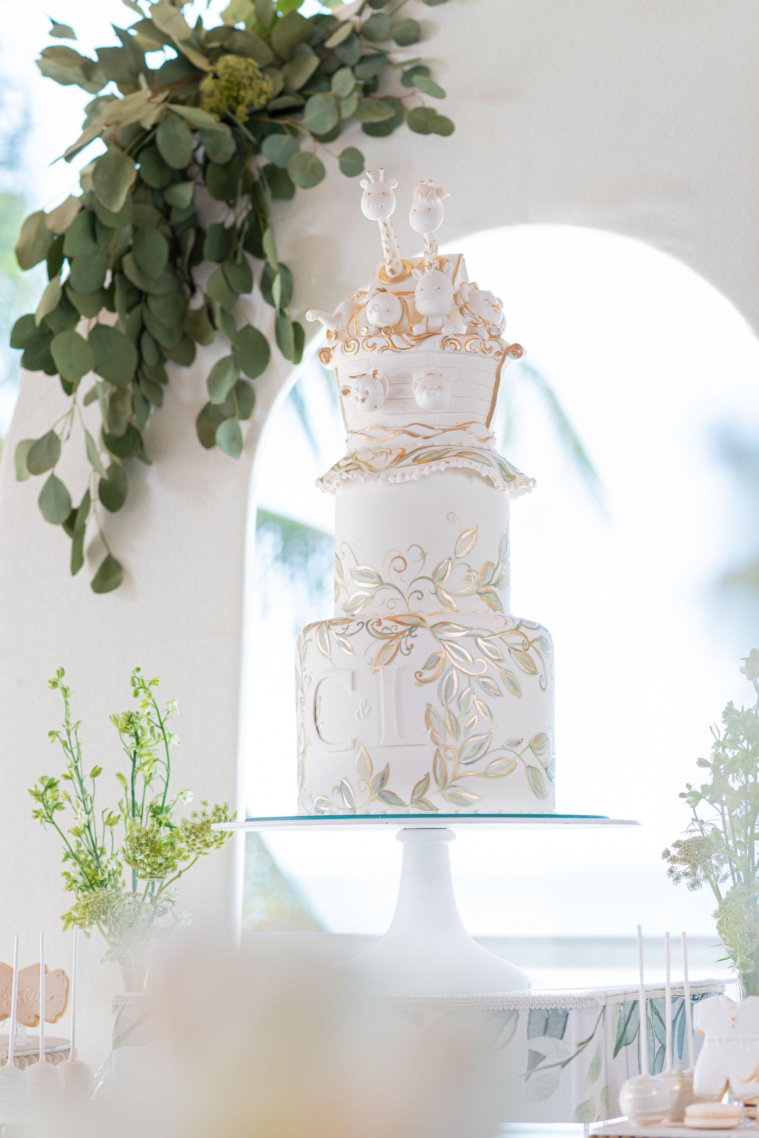 Elegant white wedding cake with delicate gold details and greenery accents displayed on a glass pedestal.