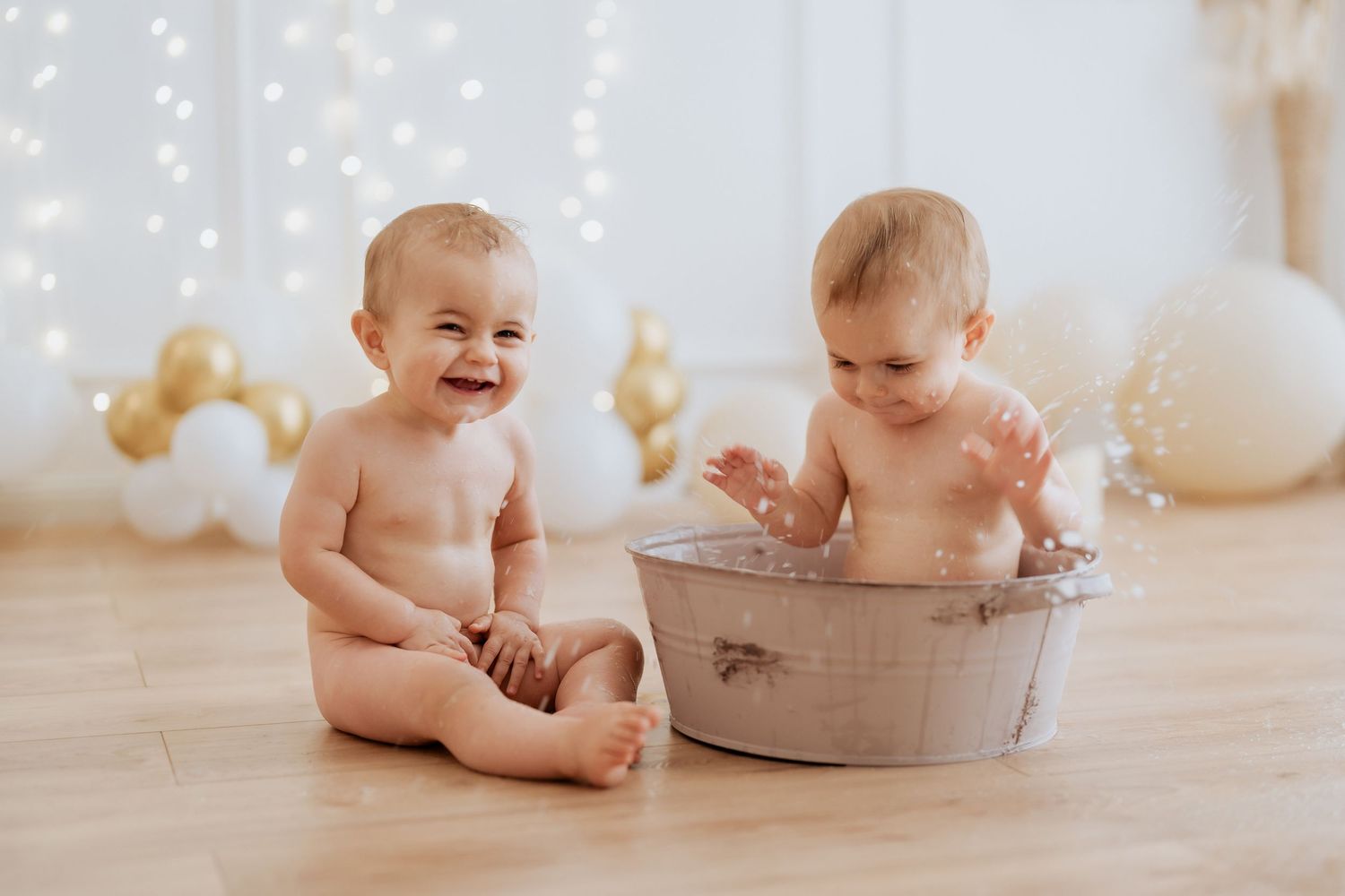 Two happy babies sitting on wooden floor with one in a vintage metal tub surrounded by festive balloons.