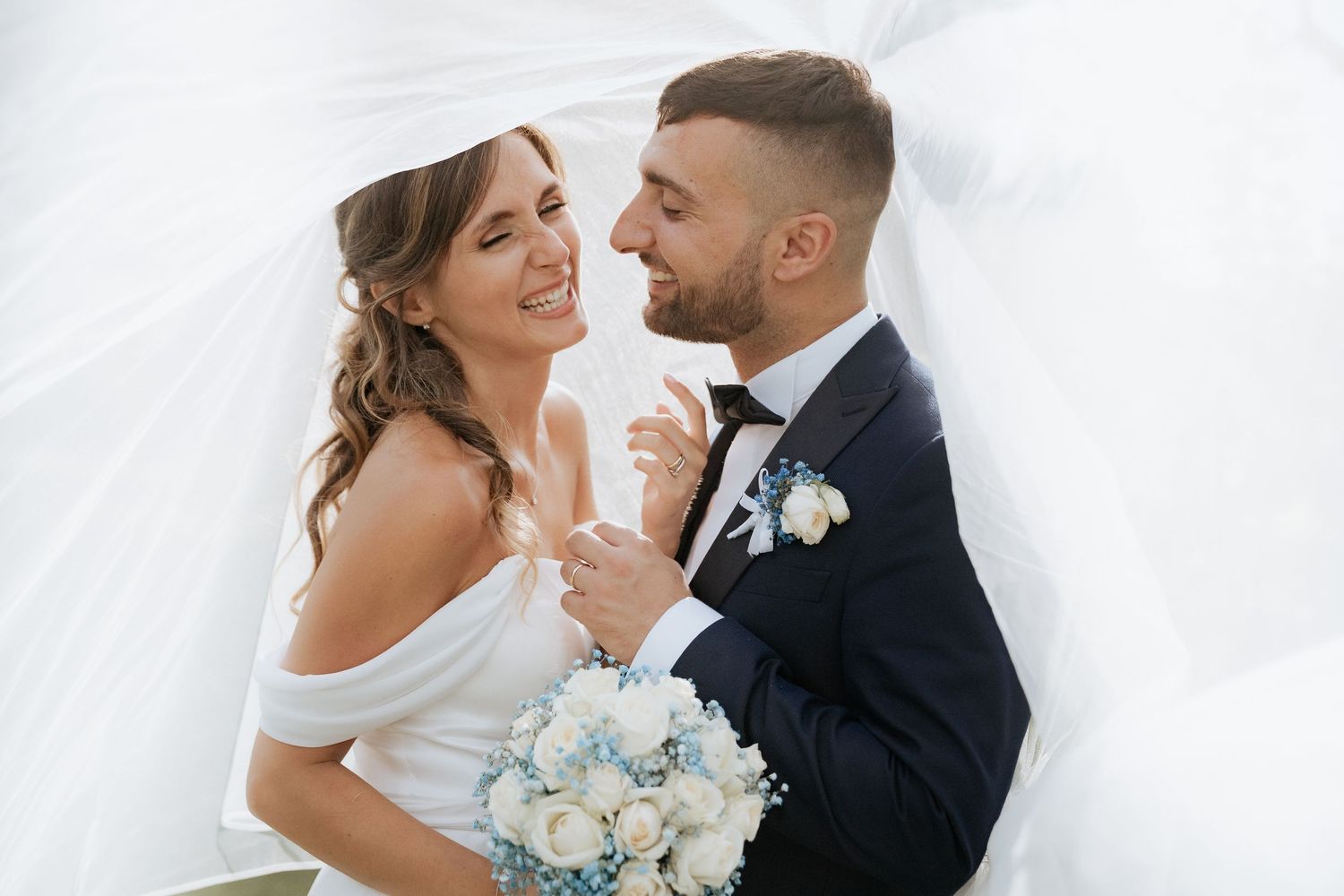 A happy couple shares a joyful moment on their wedding day while holding a bouquet of light blue flowers.