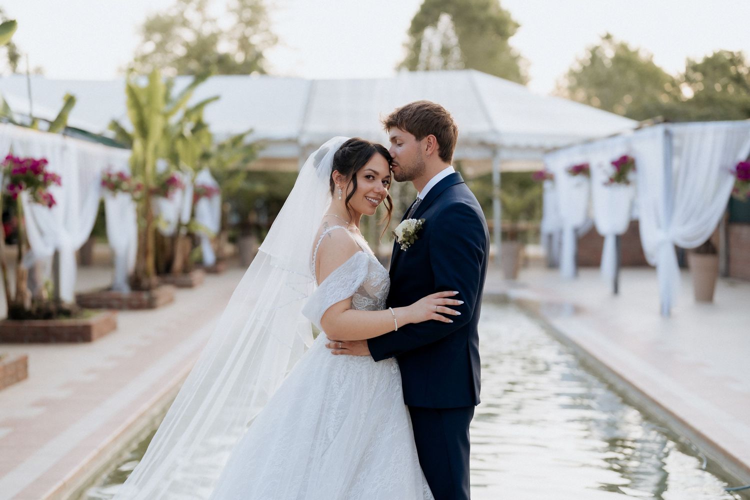 A romantic couple shares intimate moments in their wedding attire at a luxurious outdoor venue with white decor.