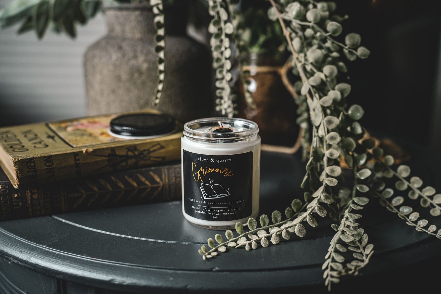Product photography of  andcrafted Grimoire scented candle jar on dark table surrounded by trailing eucalyptus stems.
