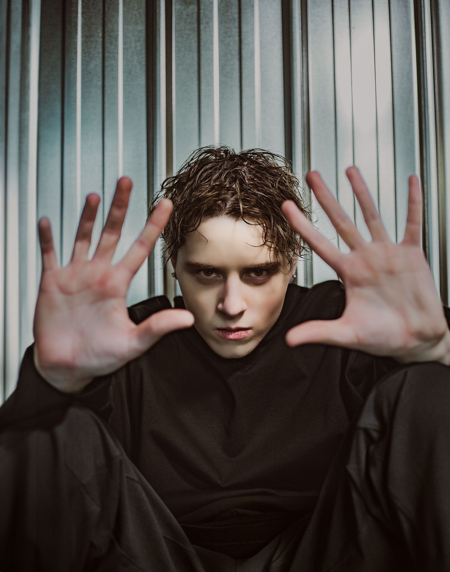 A young man with wet curly hair reaches both hands toward the camera against a corrugated metal background during model portfolio shoot.