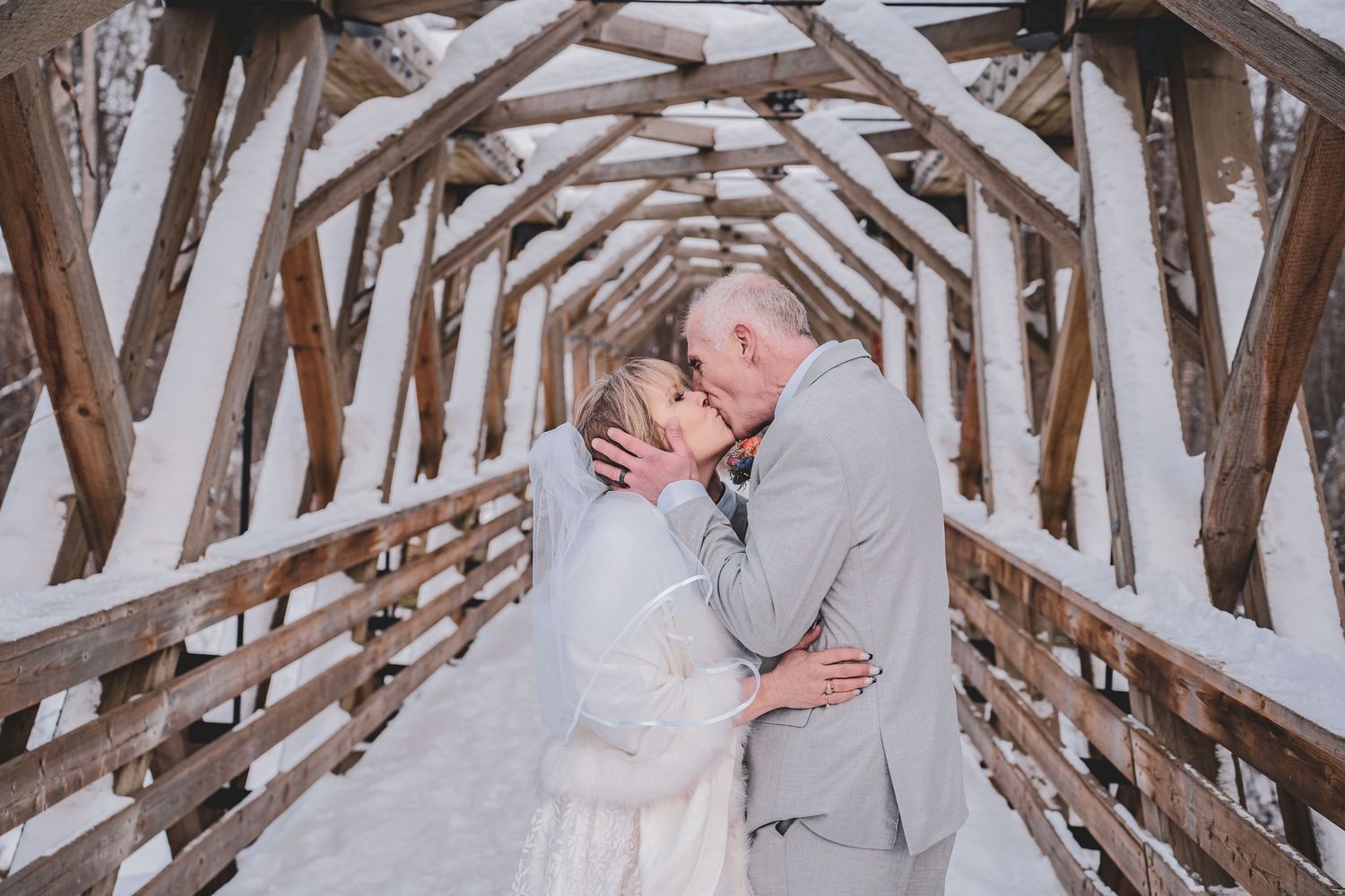 Bride and groom share a romantic kiss on a snow-covered wooden bridge in a winter wedding setting offered by Free West Photography.