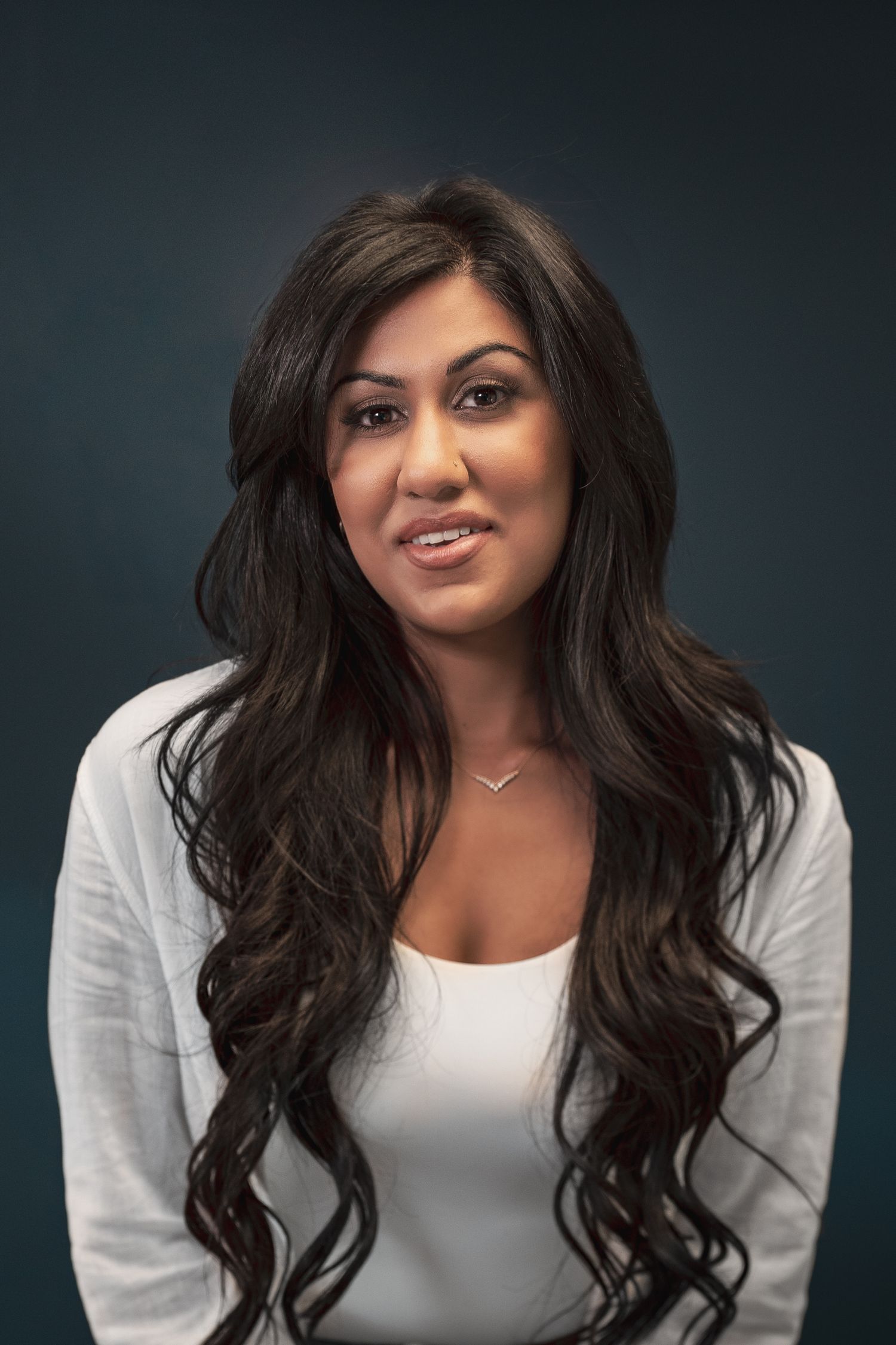 Woman with long dark wavy hair in white top smiling against dark background during her headshot photoshoot.