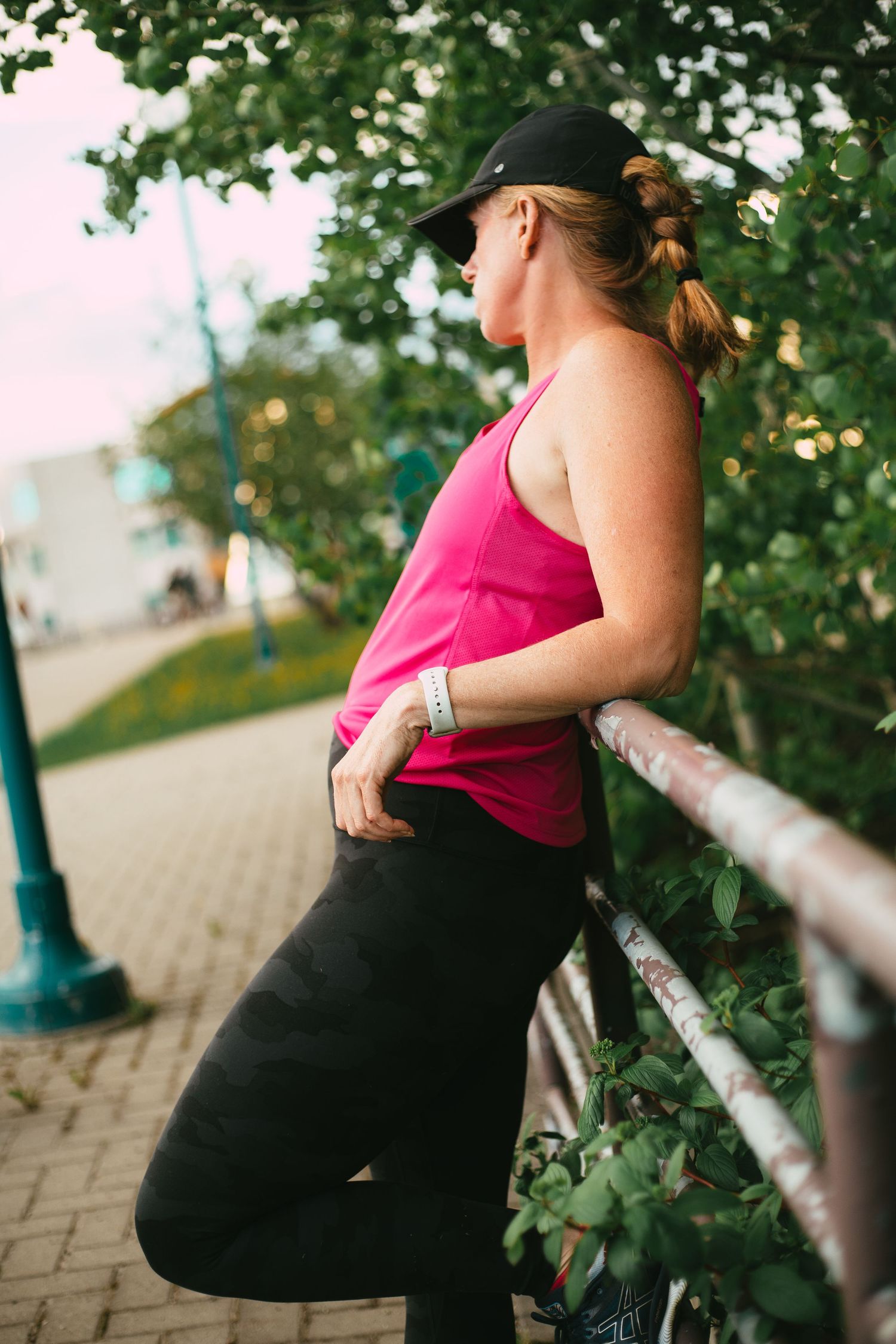 A fit woman wearing a pink tank top, black cap, and leggings rests against a metal railing outdoors for commercial business photoshoot.