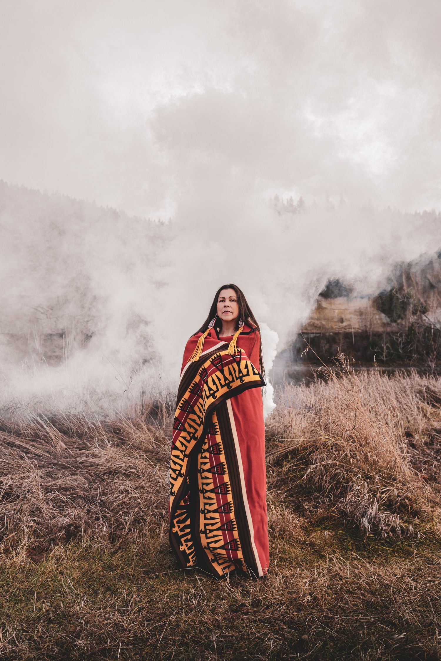 Kym Gouchie wrapped in colorful Indigenous blanket standing in misty field with foggy forest background during artist photoshoot.