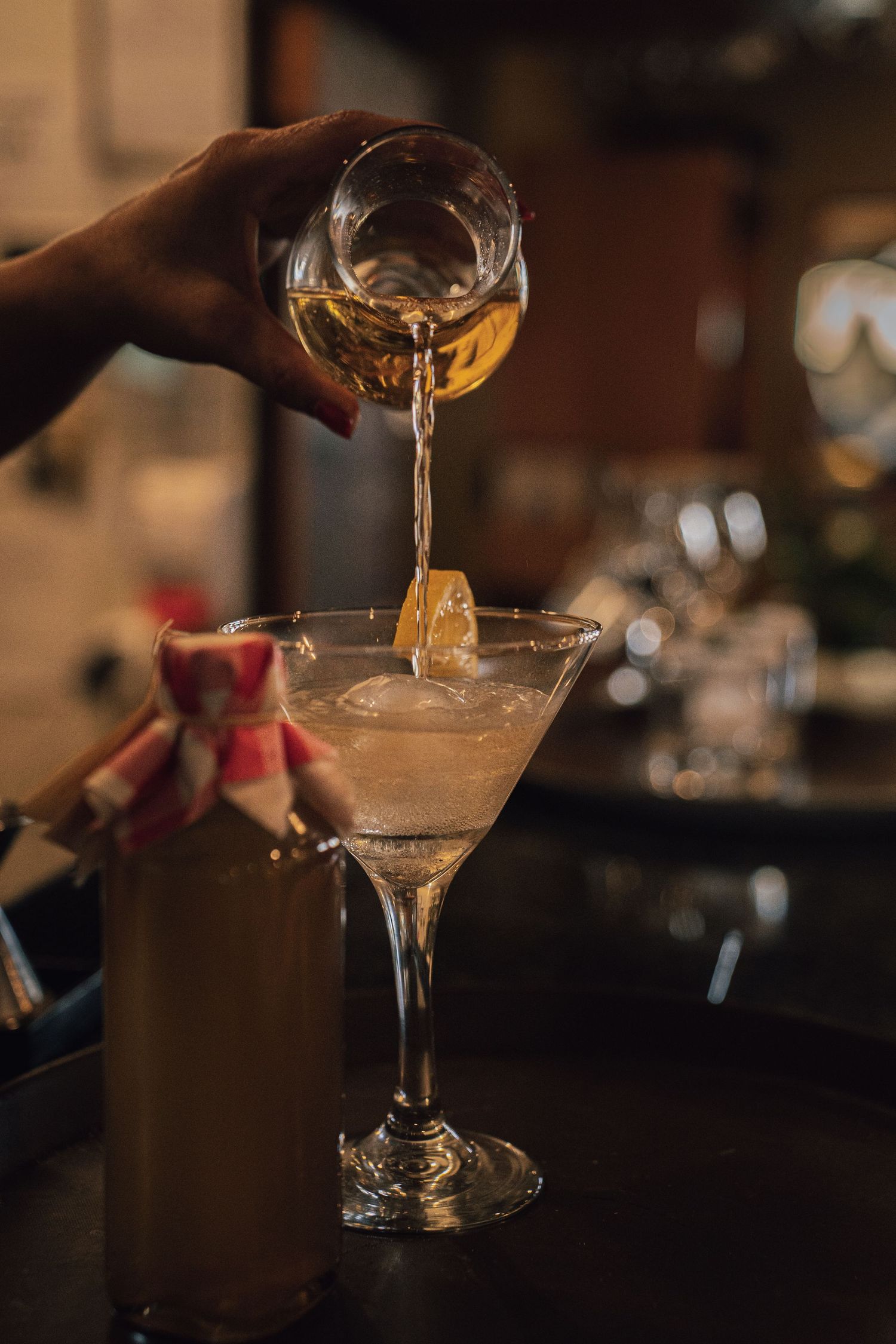 Bartender pouring golden liquor into a martini glass with ice at a dimly lit bar.