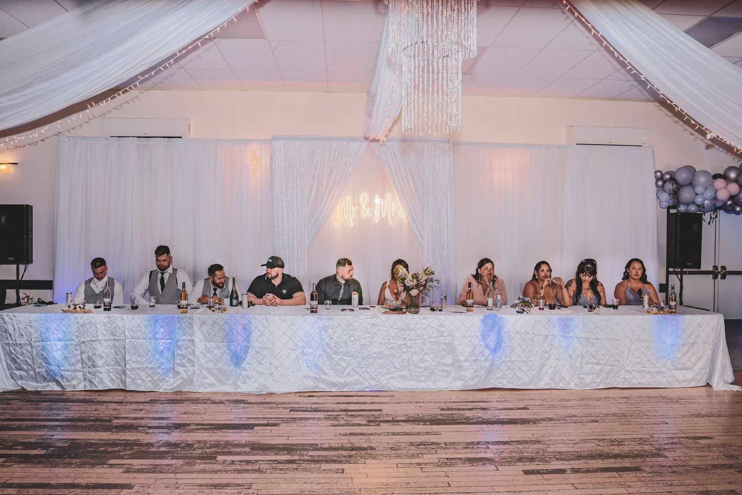 Wedding party seated at an elegantly decorated head table with white draping and chandelier backdrop.