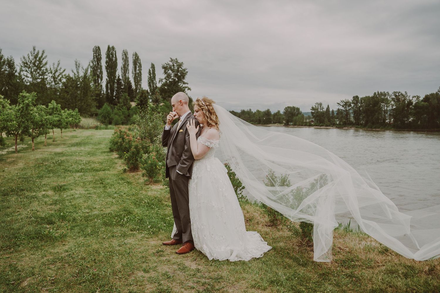 Bride and groom standing by river as long white veil billows dramatically in the wind at Northern Lights Winery, Prince George, BC.