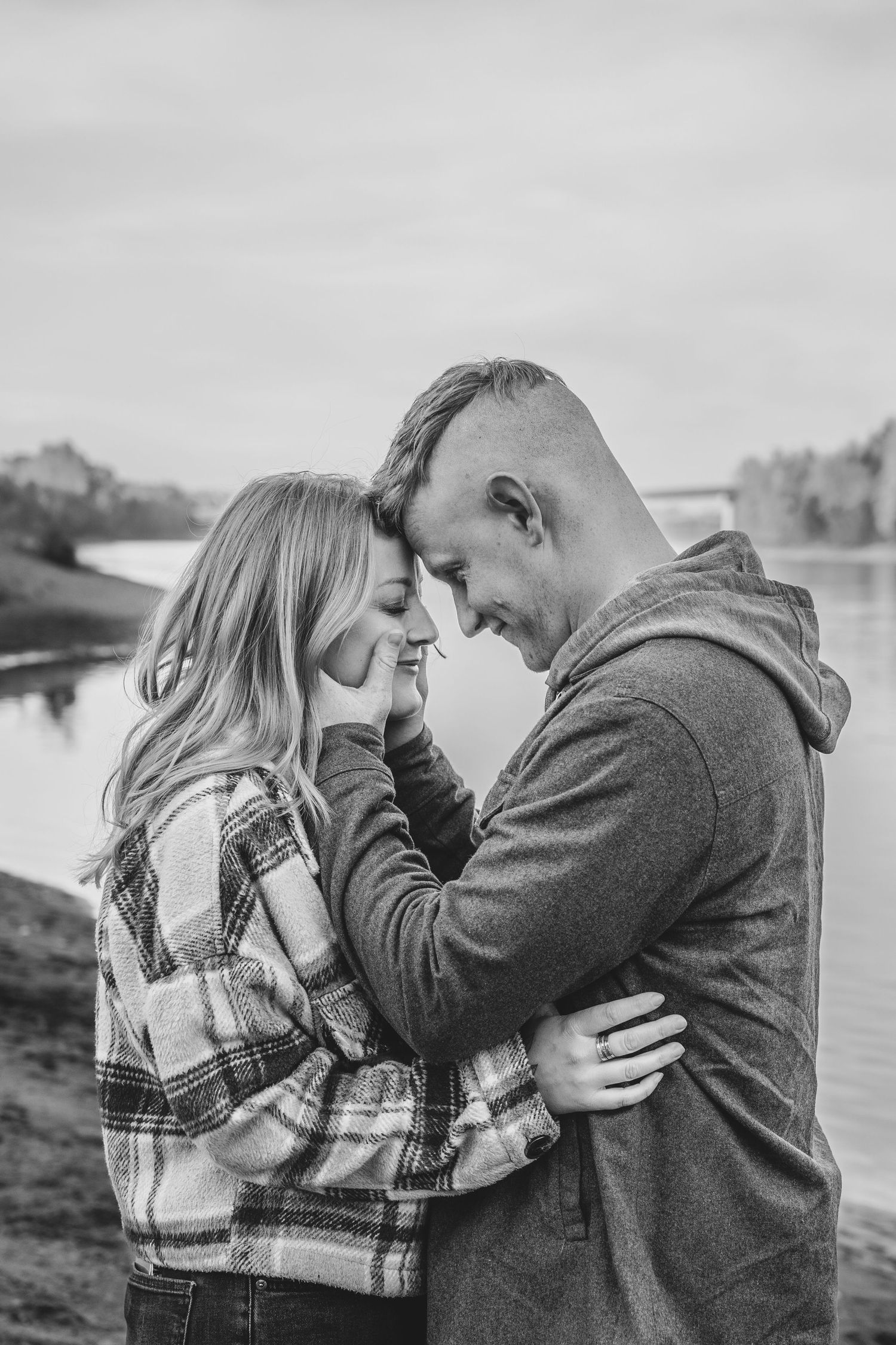 Black and white portrait of couple touching foreheads tenderly by a calm river.