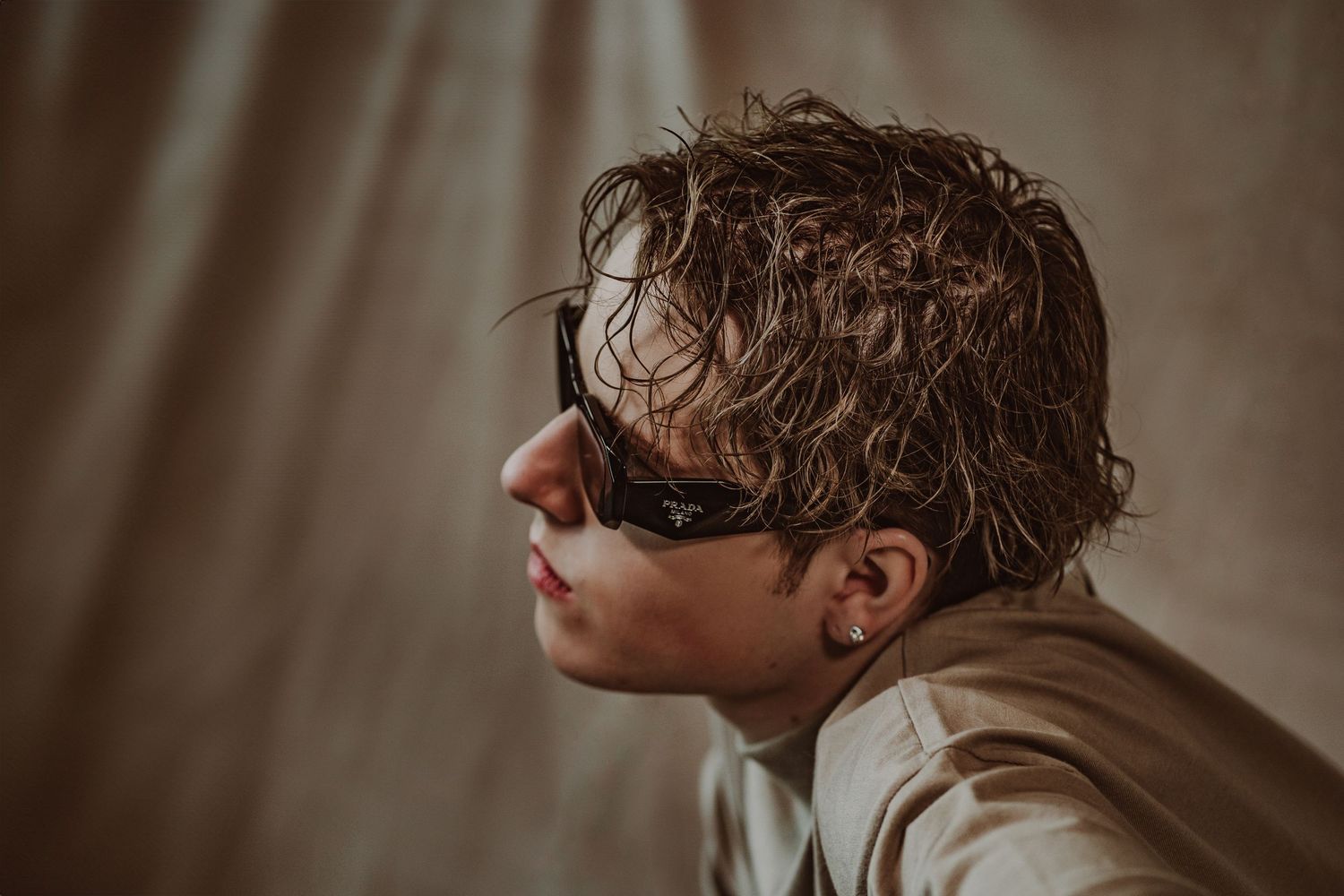 Young man with wet hair wearing black sunglasses, side profile portrait against neutral beige backdrop during a model portfolio shoot.