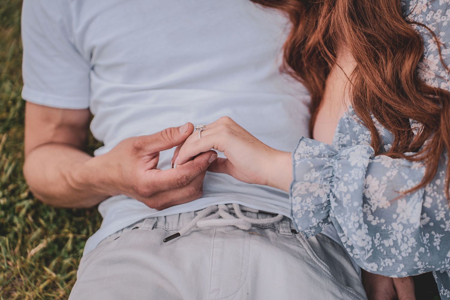 Close-up of couple holding hands showing a diamond engagement ring while sitting on grass.