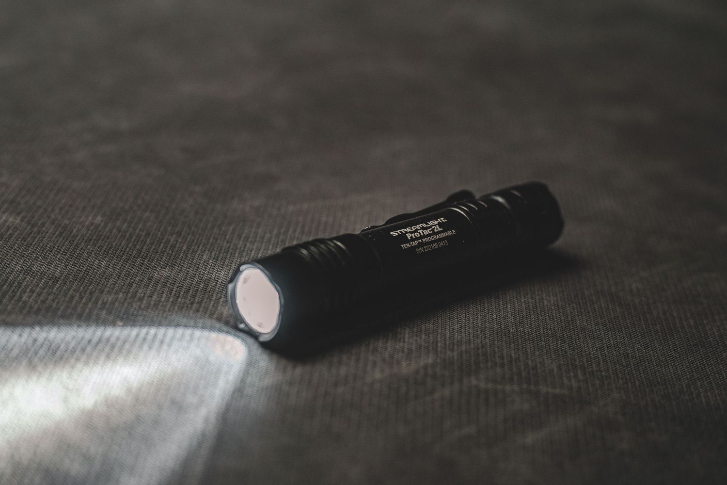Product photography of Black tactical LED flashlight emitting bright beam of light on dark fabric surface.