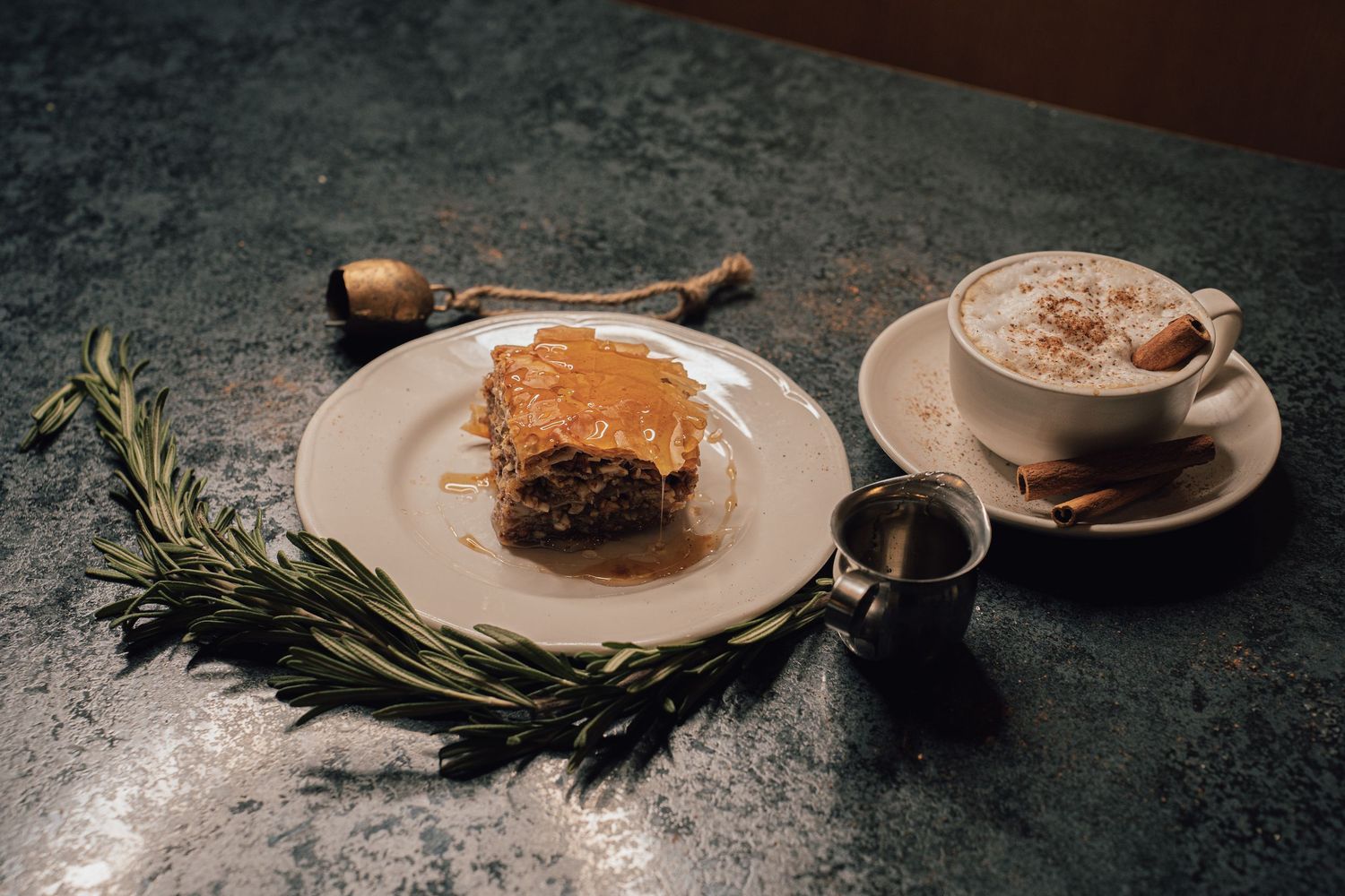 Slice of baklava on white plate beside a cappuccino with rosemary garnish on marble surface.