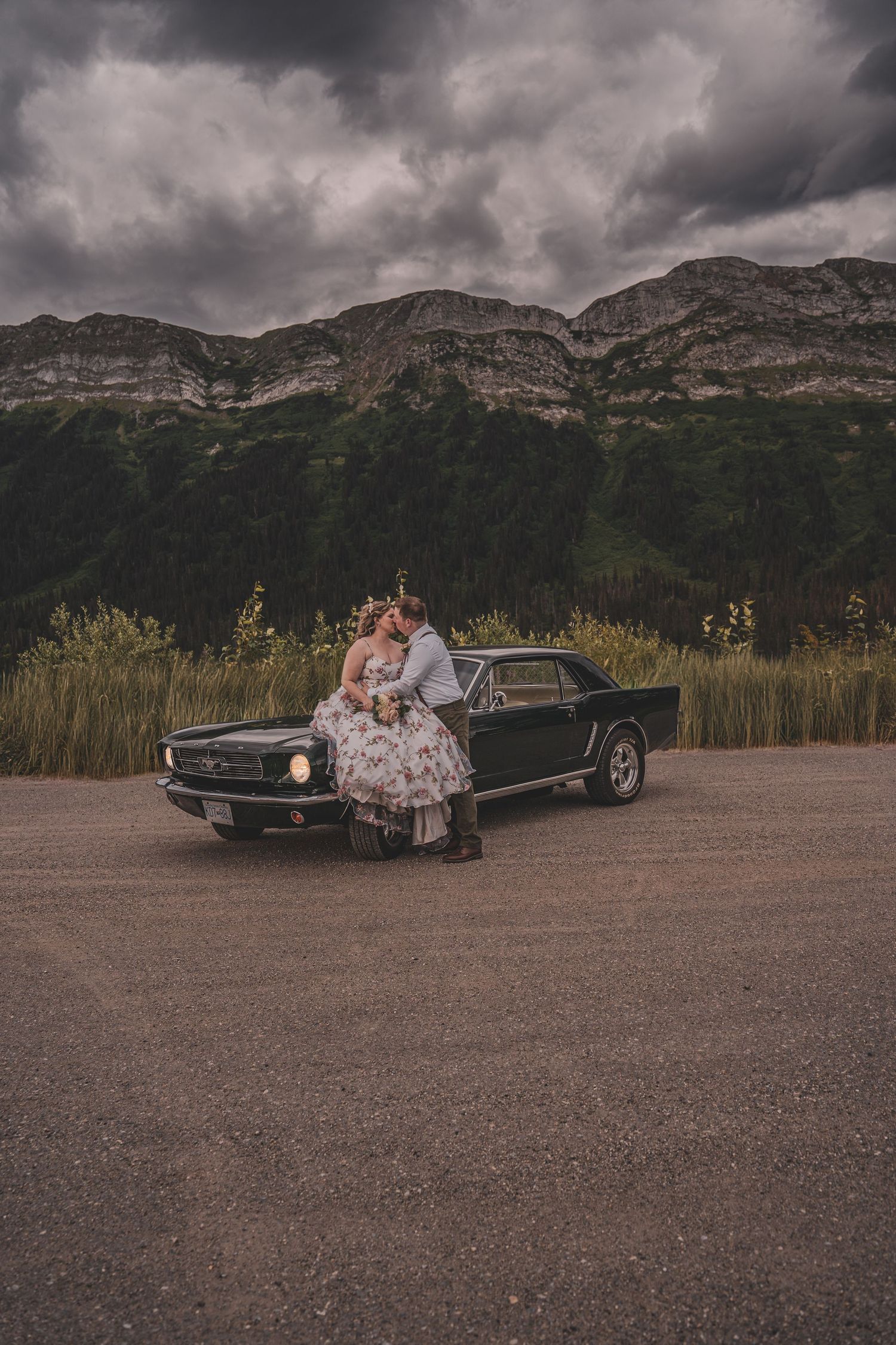 Bride and groom kiss leaning against a classic black vintage Mustang with dramatic mountain backdrop.