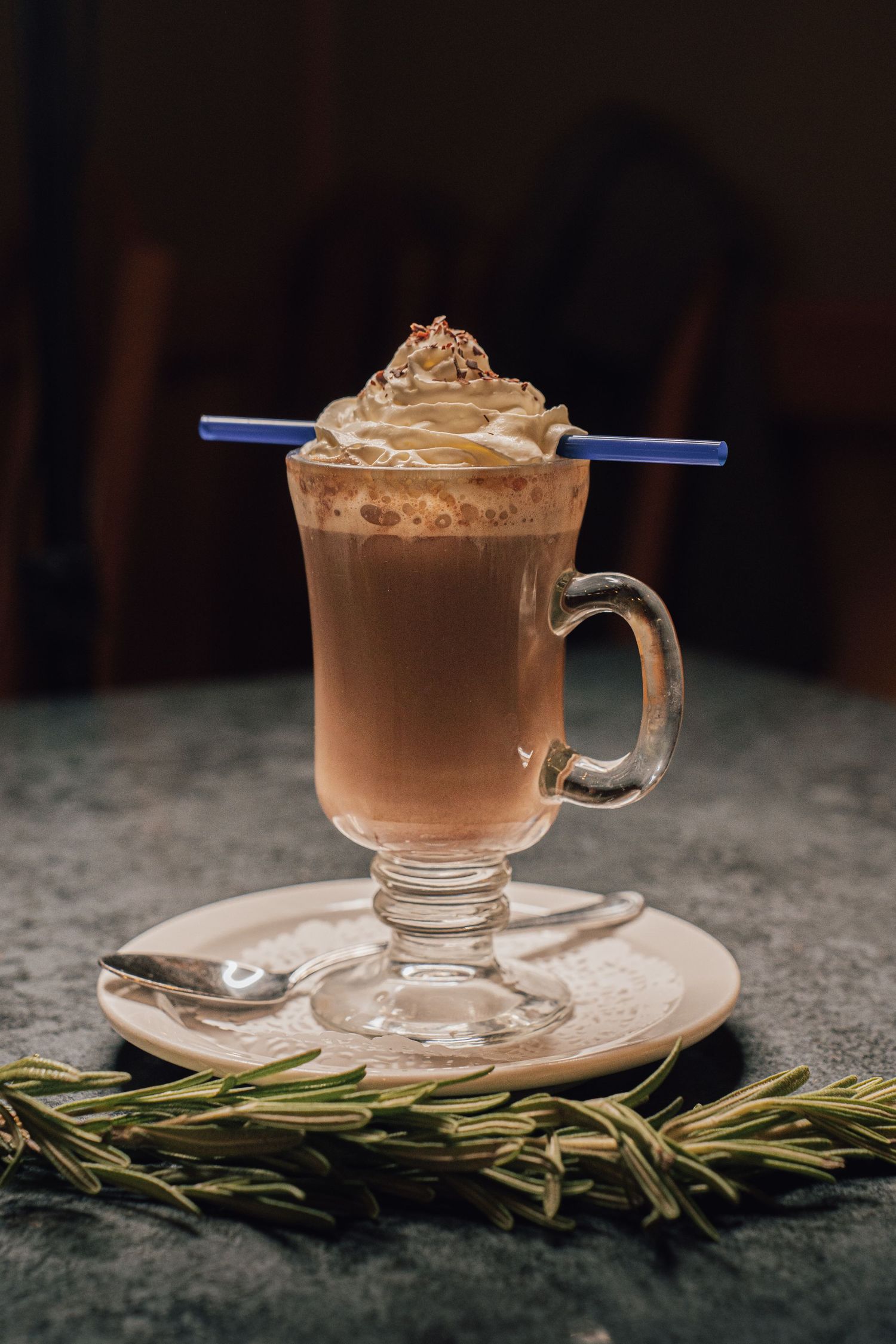 Tall glass mug of creamy coffee topped with whipped cream and a blue straw beside rosemary.