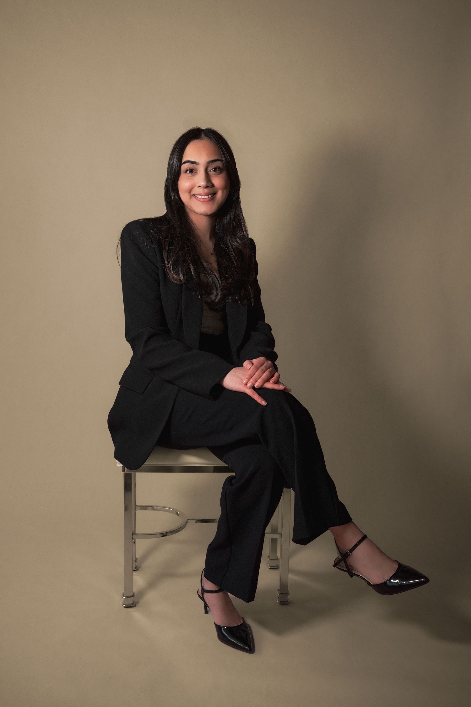 Smiling young woman in black pantsuit seated on chair against neutral background during her real estate headshot photoshoot.