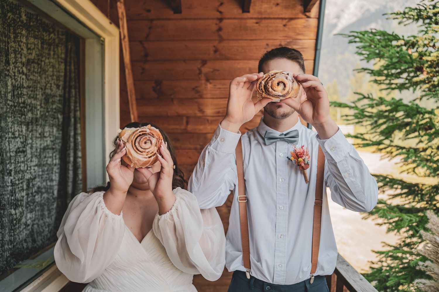 Playful bride and groom hold cinnamon rolls over their eyes on a rustic cabin porch with pine trees.