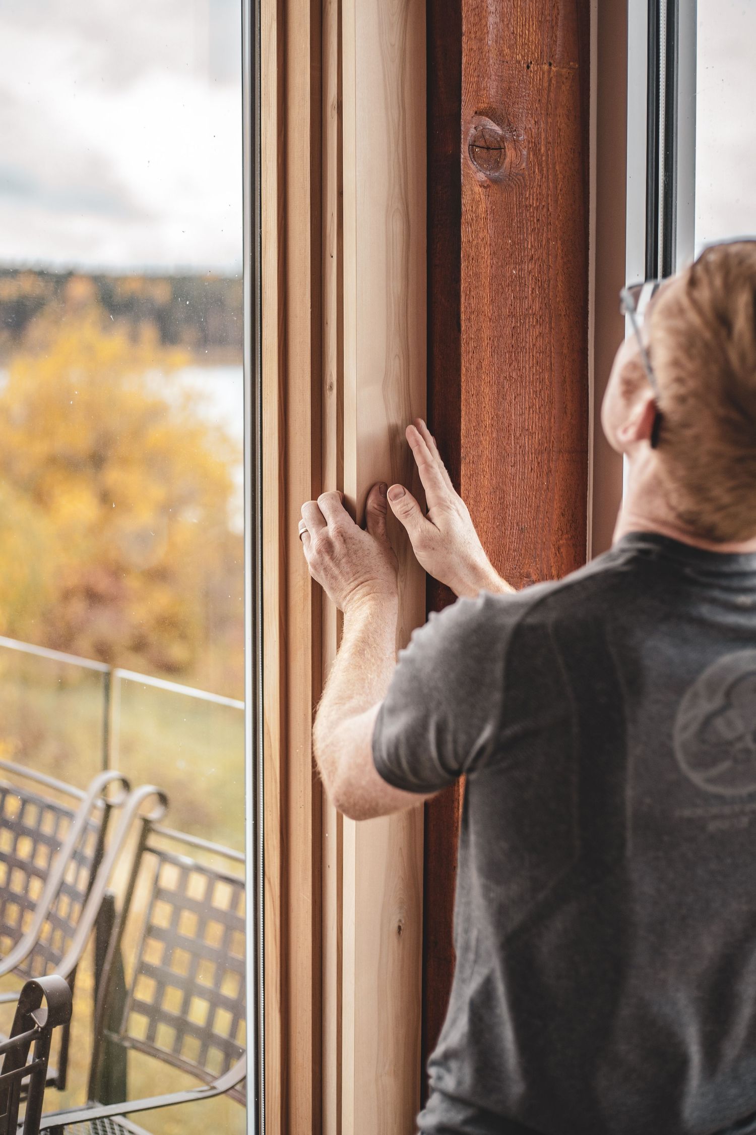 Carpenter installing wooden window trim with scenic autumn lake view visible through glass.