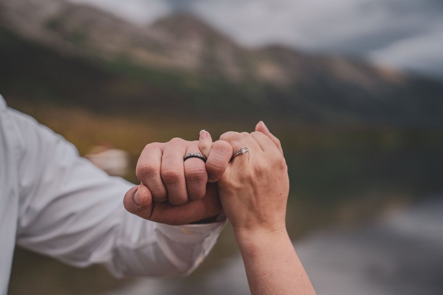 Couple holds hands showing matching wedding bands with a scenic mountain lake in the background.