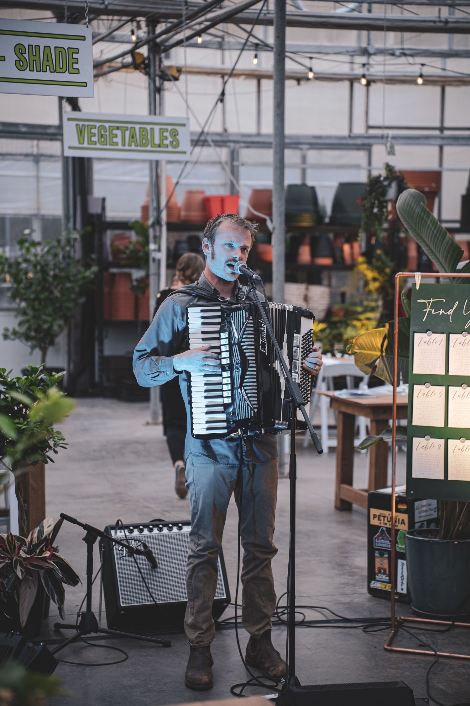A musician plays accordion and sings into a microphone inside a plant-filled greenhouse venue.
