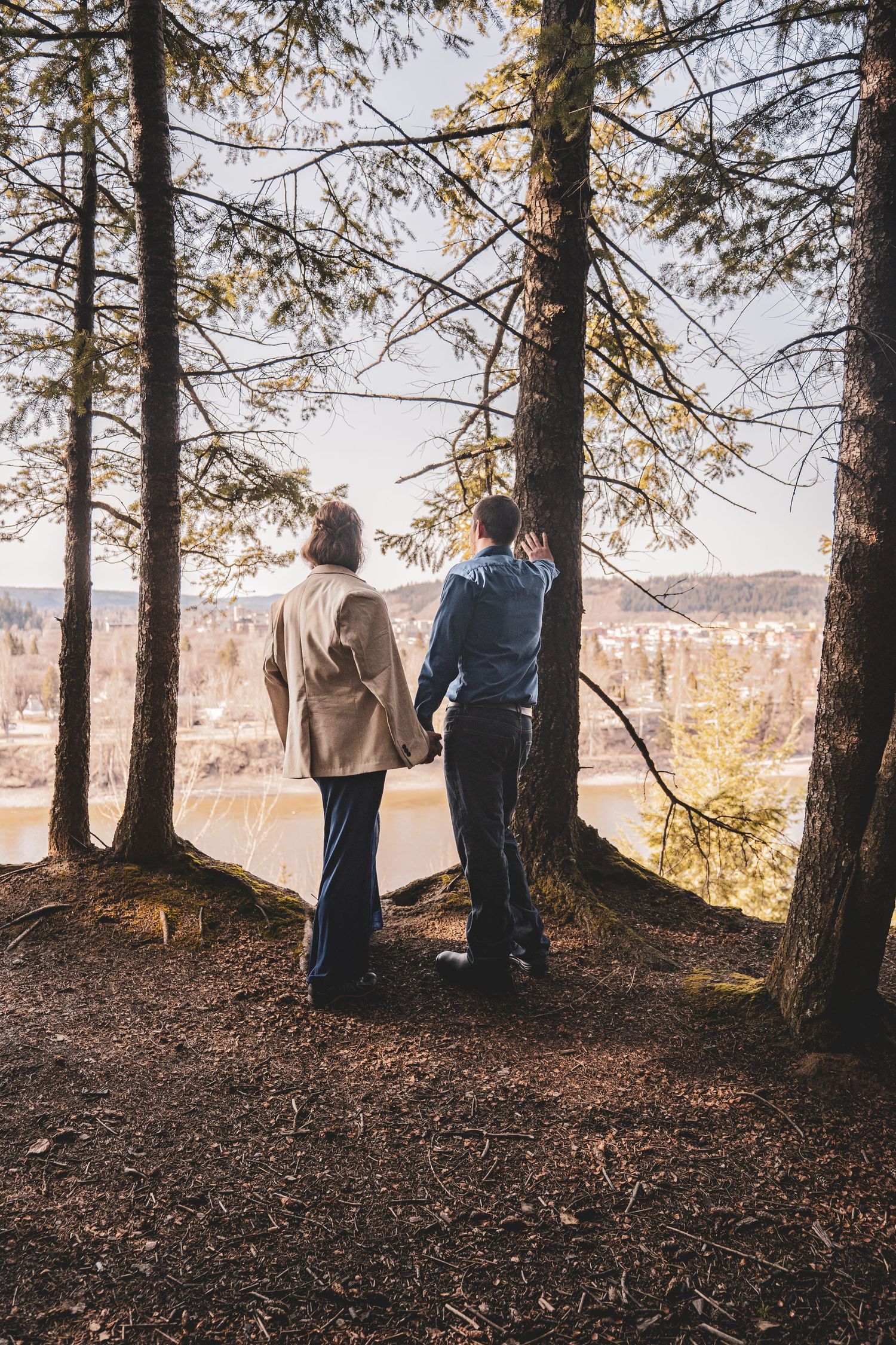 Couple holding hands standing among pine trees overlooking scenic river valley landscape.