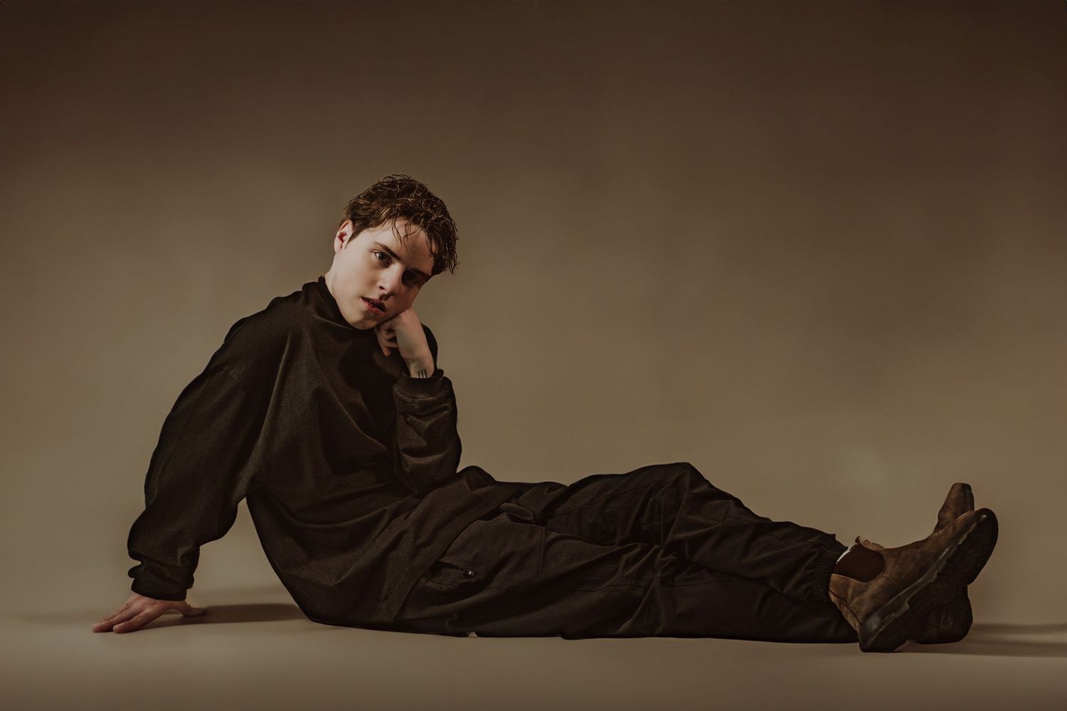 young artist in black oversized outfit seated on floor in moody studio portrait photography.